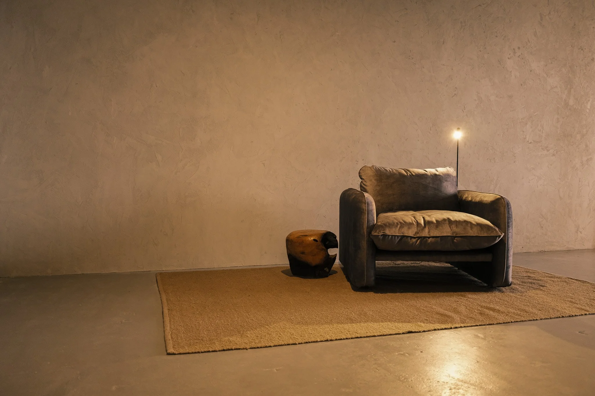 A brown armchair with a cushion, a small wooden side table, and a floor lamp against a textured beige wall.