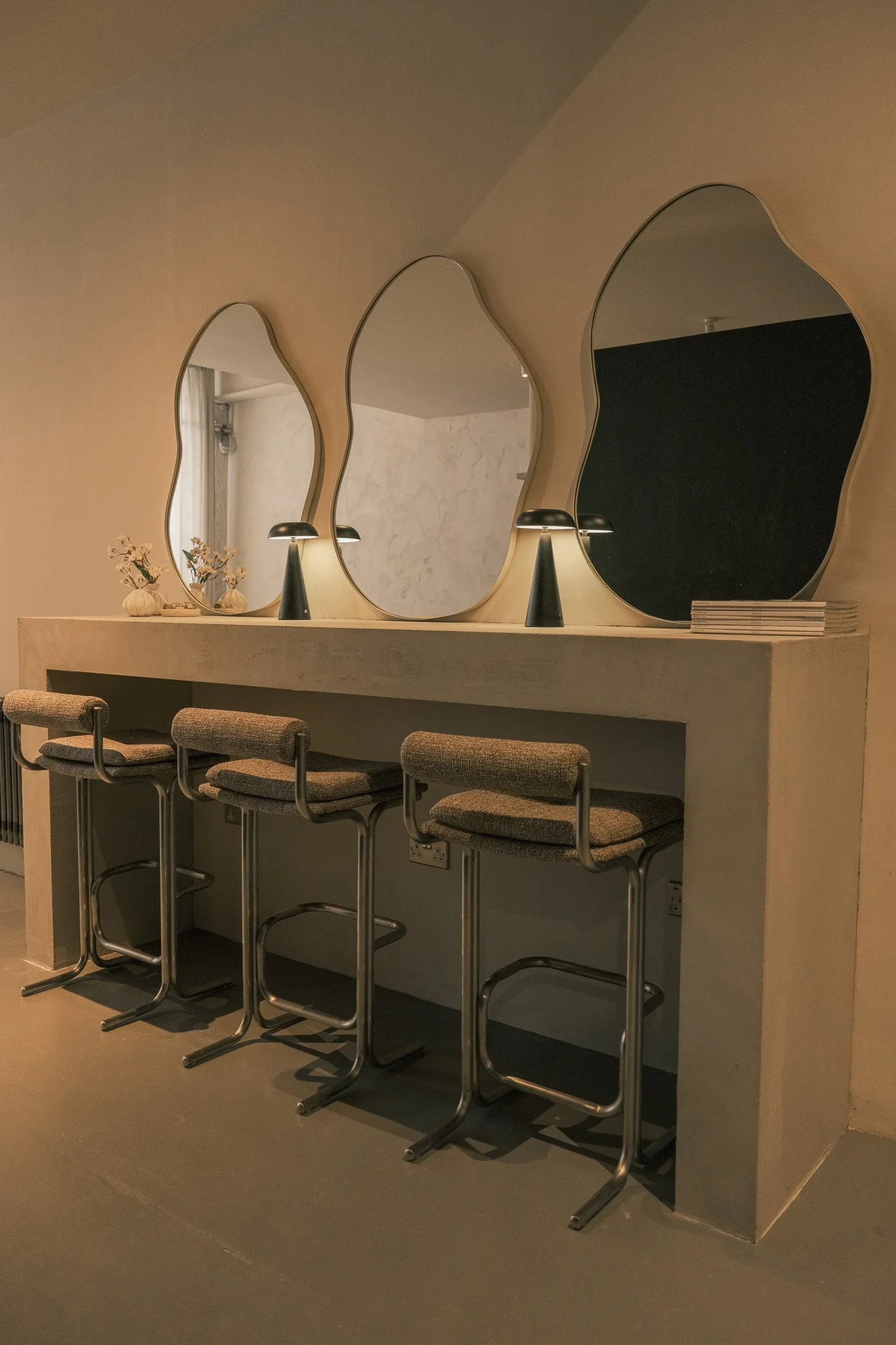 Four bar stools with cushioned seats beneath a long console table with three irregular-shaped mirrors and small lamps.