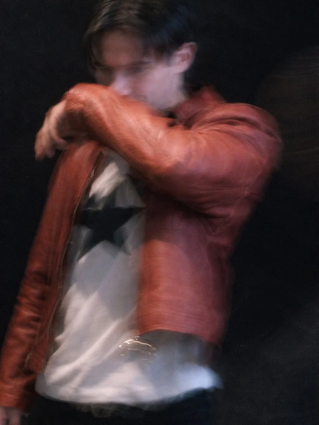 A young man with dark hair wearing a red leather jacket and a white t-shirt with black star pattern, standing against a dark background, with his arms crossed over his face.