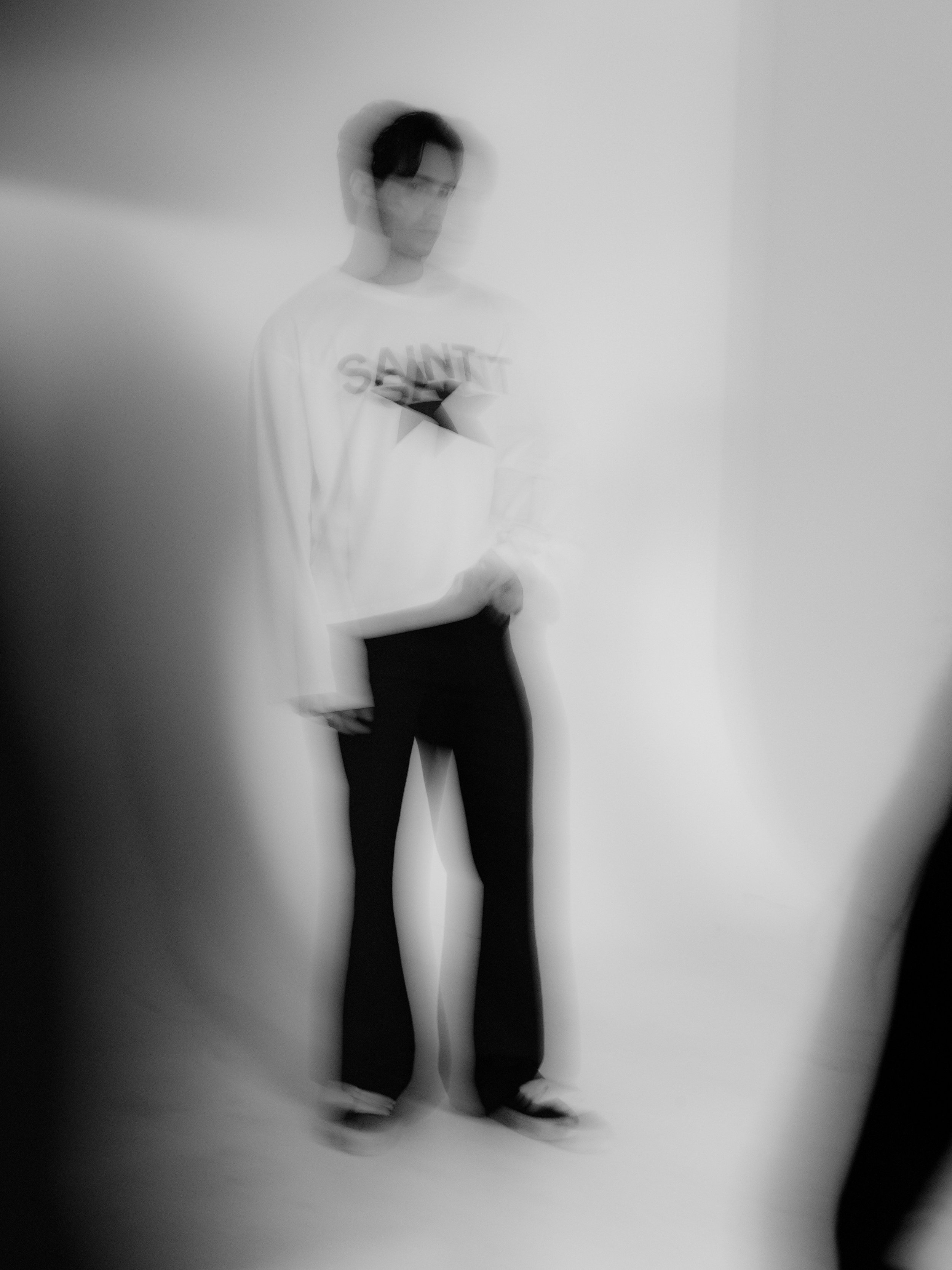Blurry black-and-white photo of a person standing against a plain white background, holding a smartphone, wearing a long-sleeve shirt with the word 'SAINT' on it, and black pants.