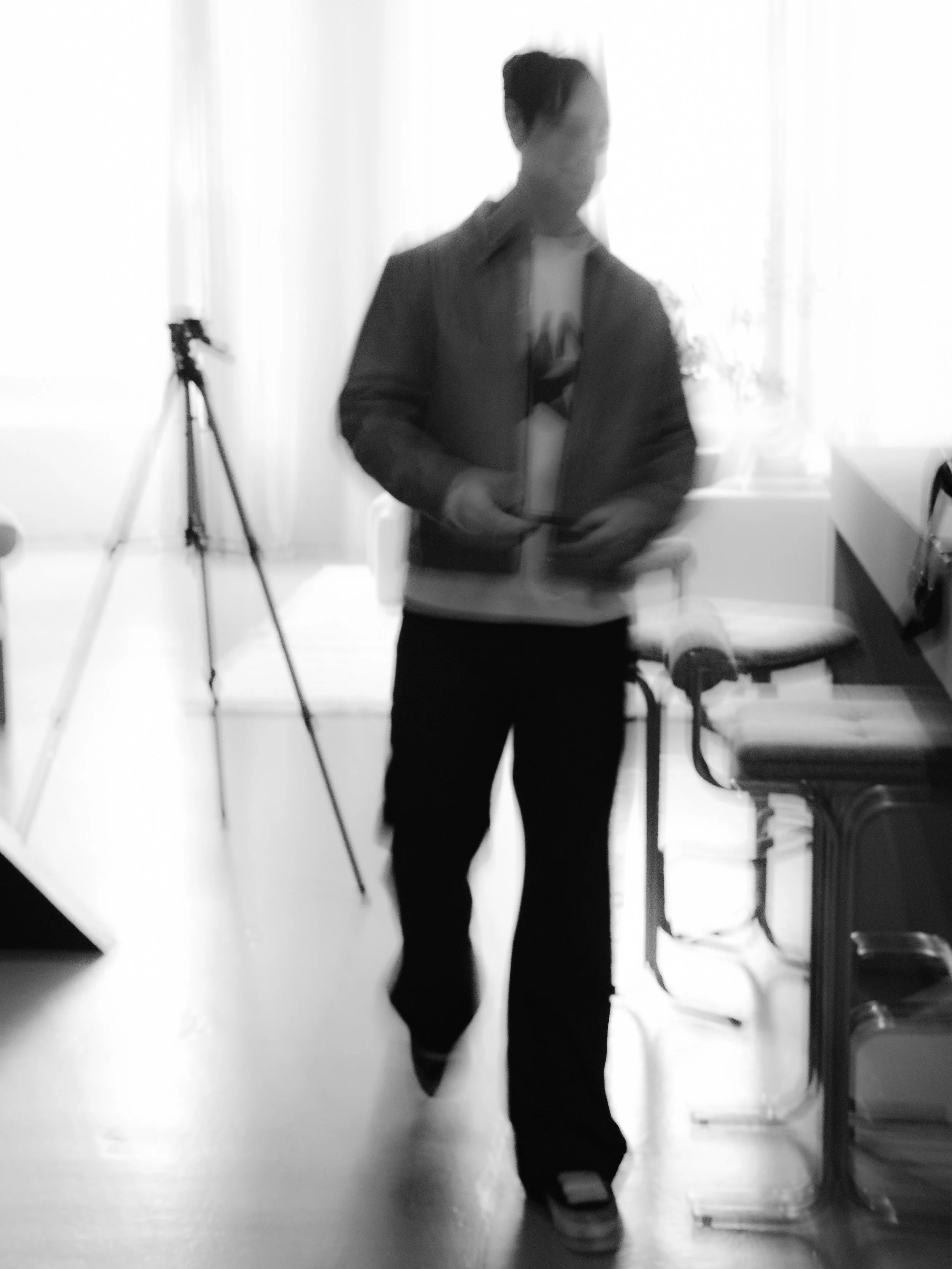 Blurry black-and-white photo of a young man in a casual jacket and pants walking in a room with a tripod, chair, and window in the background.