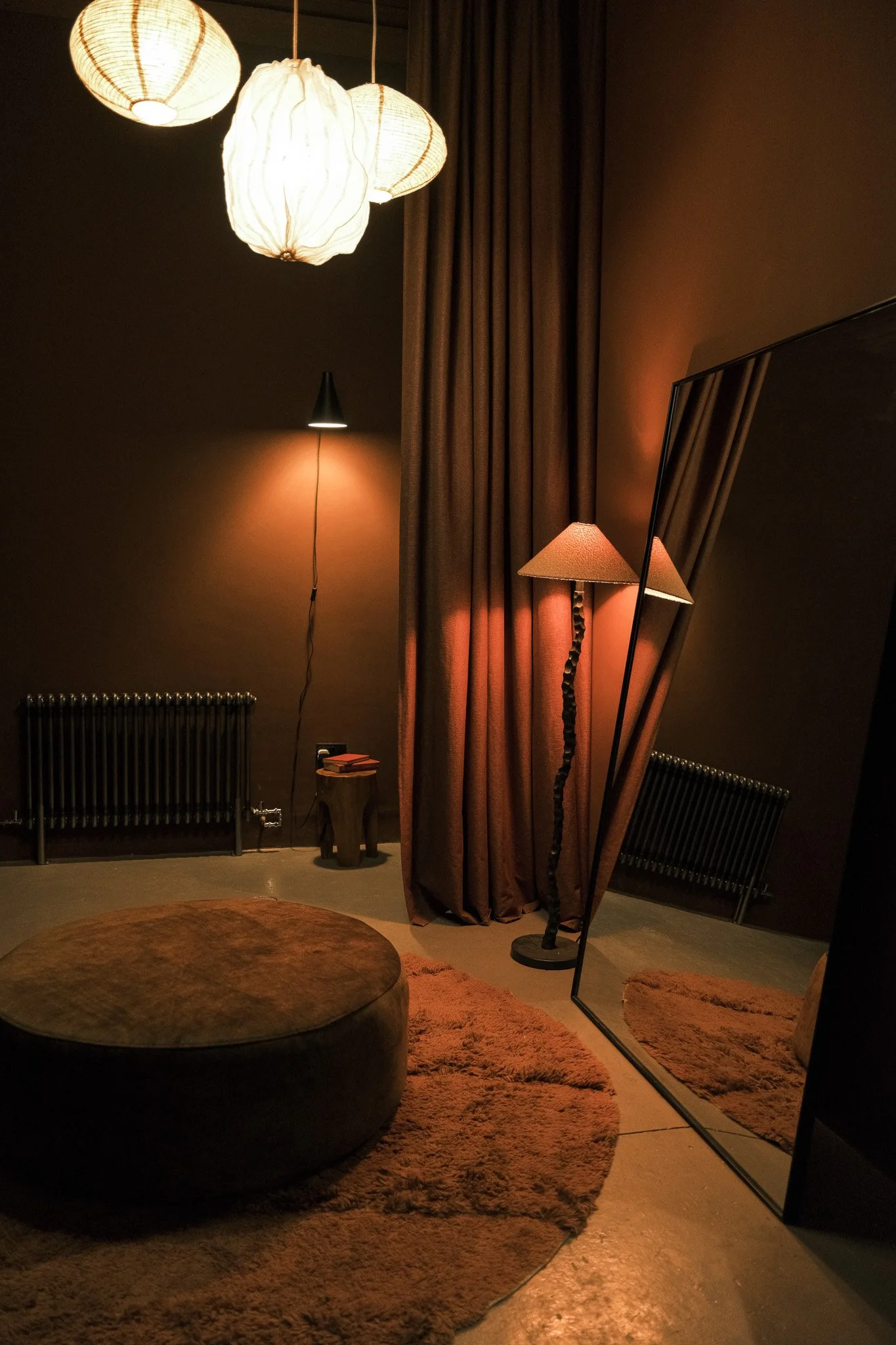 A dimly lit room with hanging paper lanterns, a floor lamp with a twisted stand, a large mirror, brown curtains, a small side table with books, a round brown ottoman, a textured rug, and radiators visible in the background.
