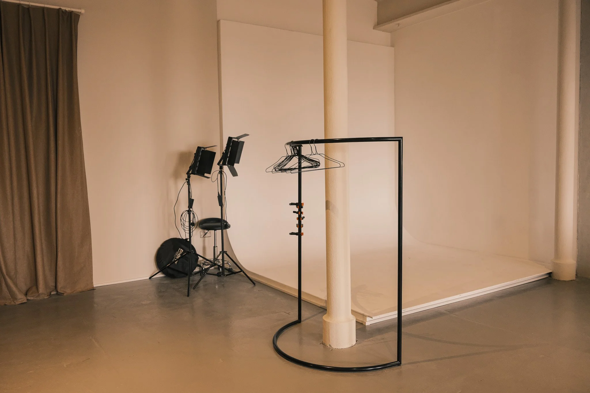 Empty photography studio with a white backdrop, studio lights on the left, a black clothing rack in the foreground, and beige curtains on the side.
