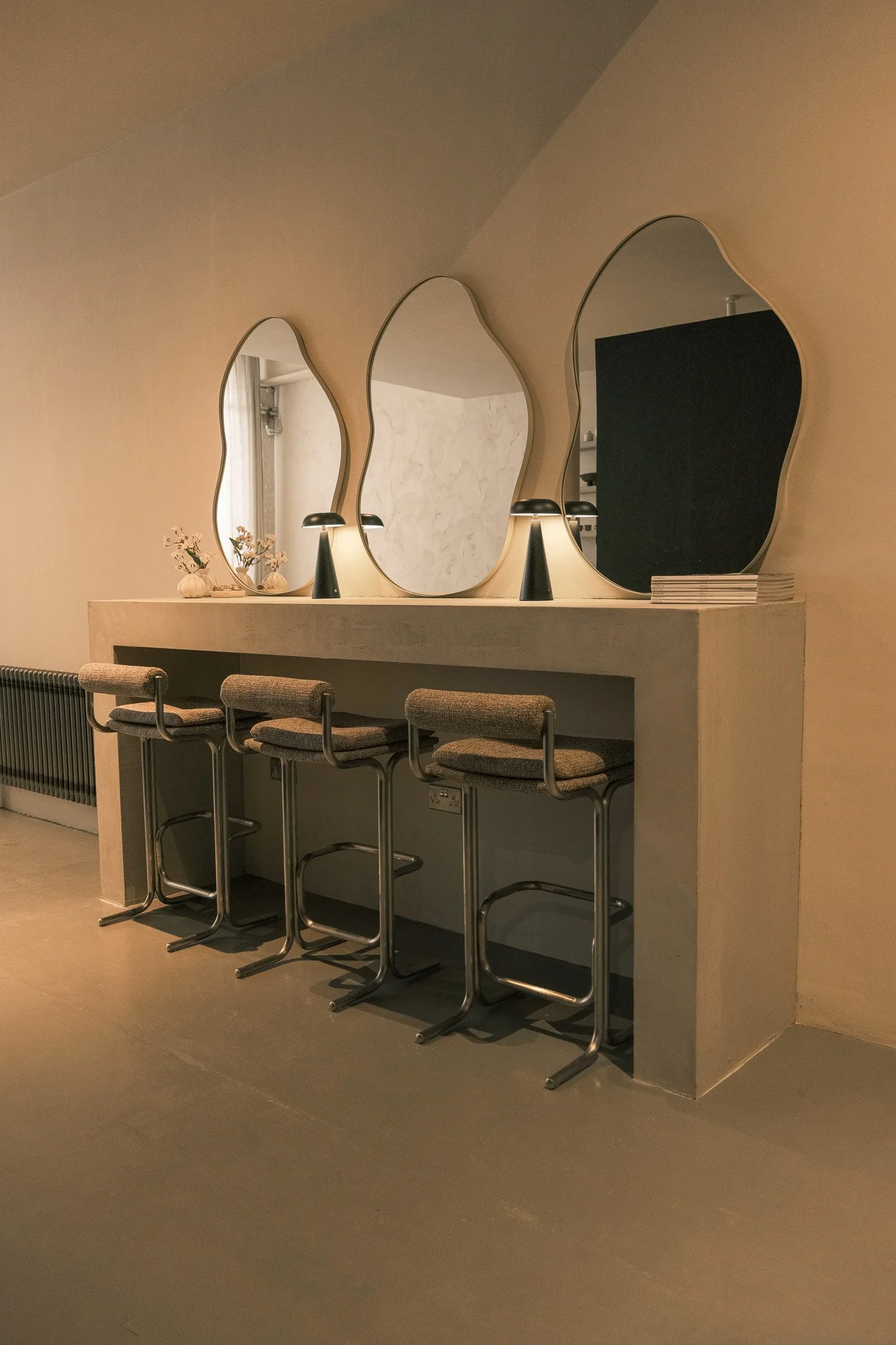 A minimalist interior with three irregularly shaped mirrors on a counter, lit by two small black table lamps, with four bar stools underneath and a stack of magazines on the right.