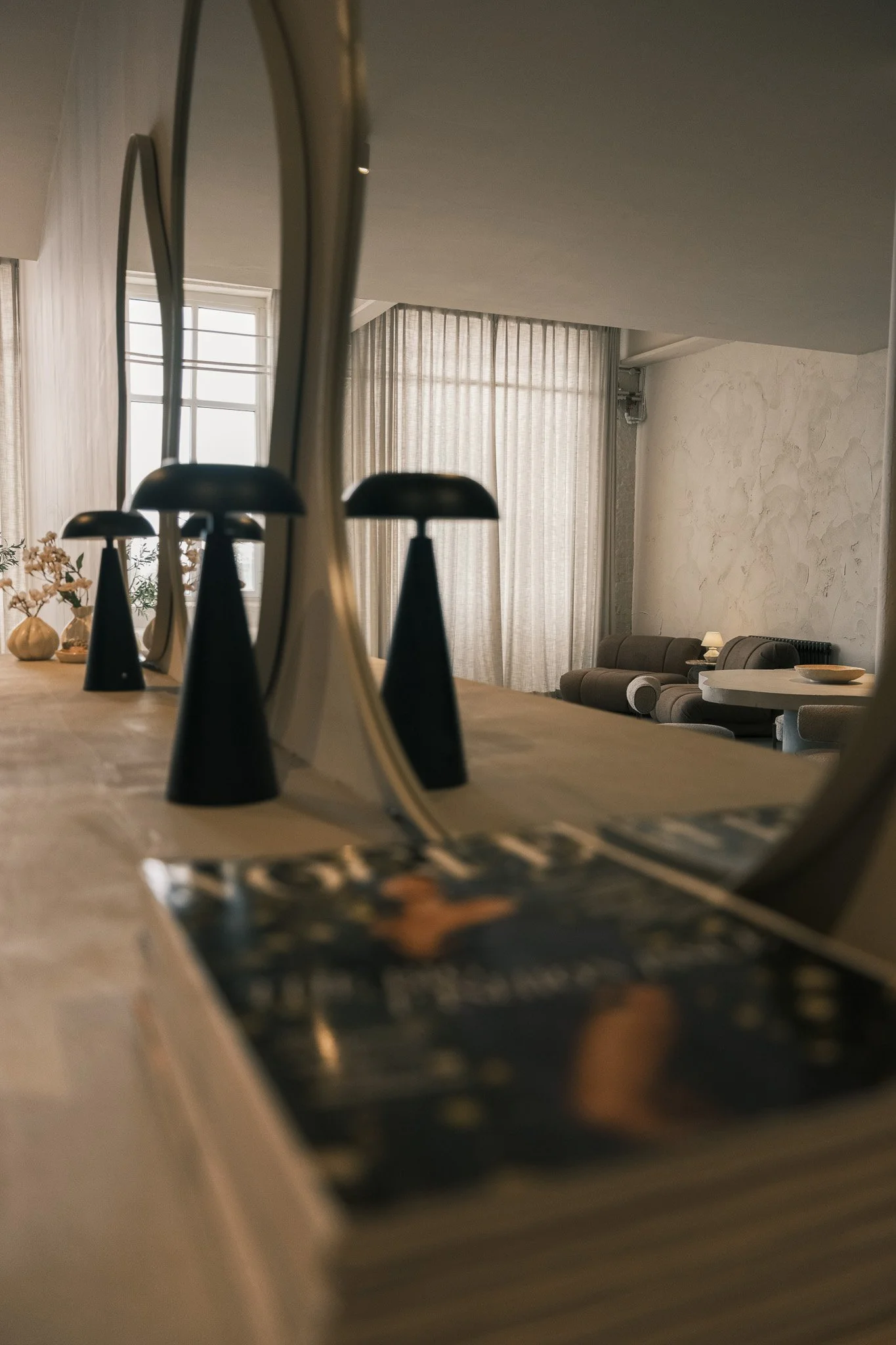 Living room with large window, beige curtains, and a seating area with a sofa and pillows, as well as a table lamp.