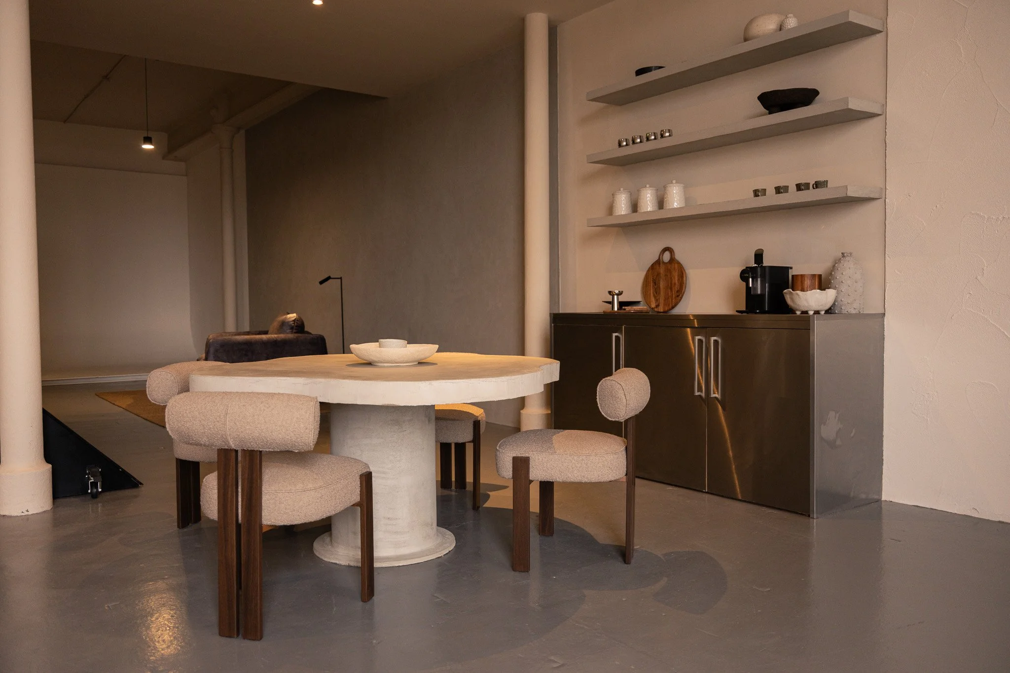 Modern dining area with a round stone table and four upholstered chairs, wicker-like on the outside, in a minimalist style. Behind the table is a cabinet with white shelves holding decorative bowls and jars, and a coffee machine on top. The room feat