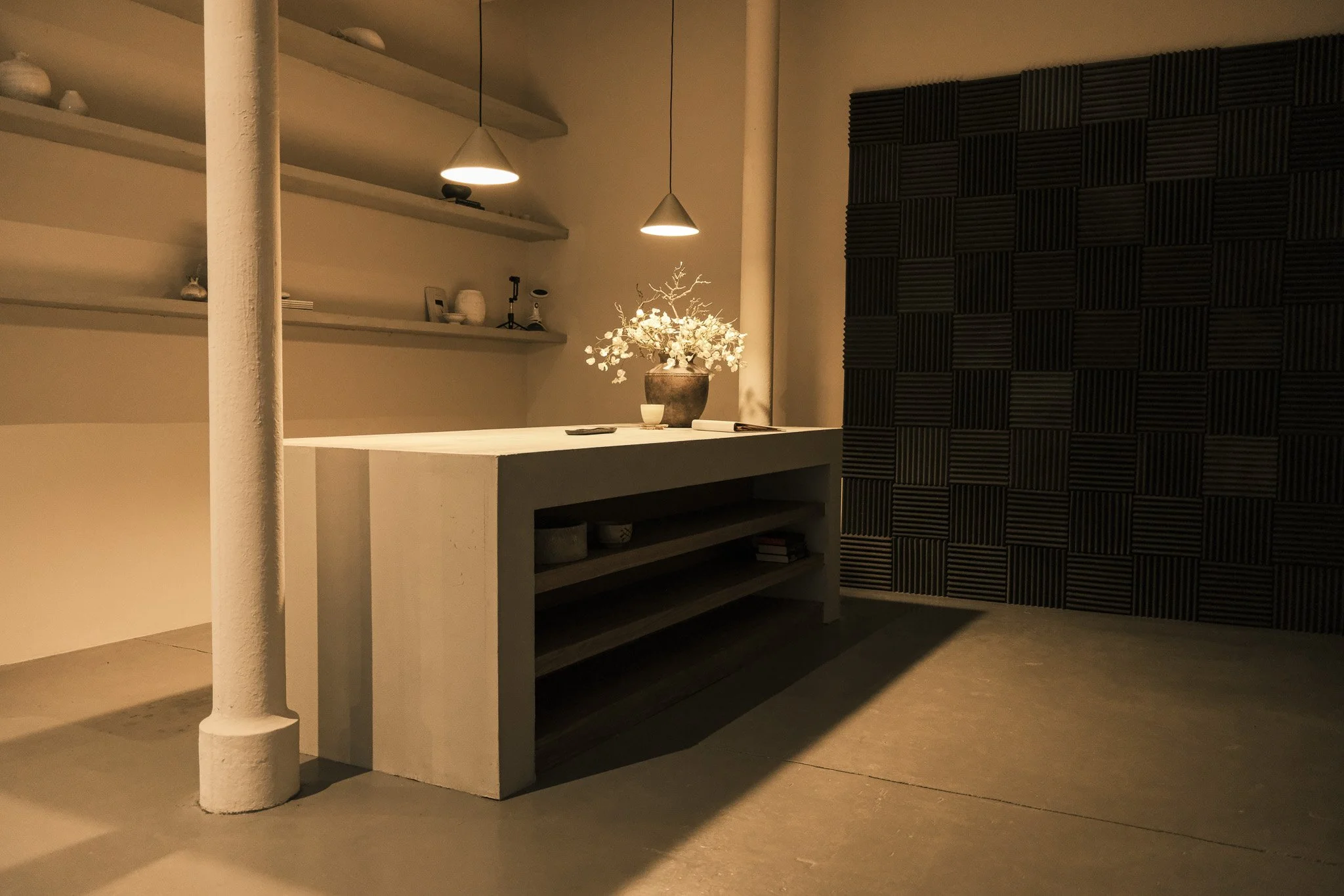 Minimalist interior space with a white table, decorative items, and a vase with white flowers under hanging pendant lights, contrasted by a black acoustic wall panel.