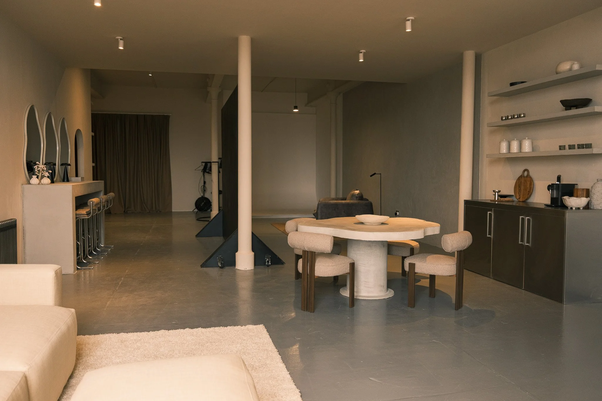 Modern, minimalist living space with a round dining table and four beige upholstered chairs, open shelving with decorative items, a gray sofa, and a cozy armchair in the background.
