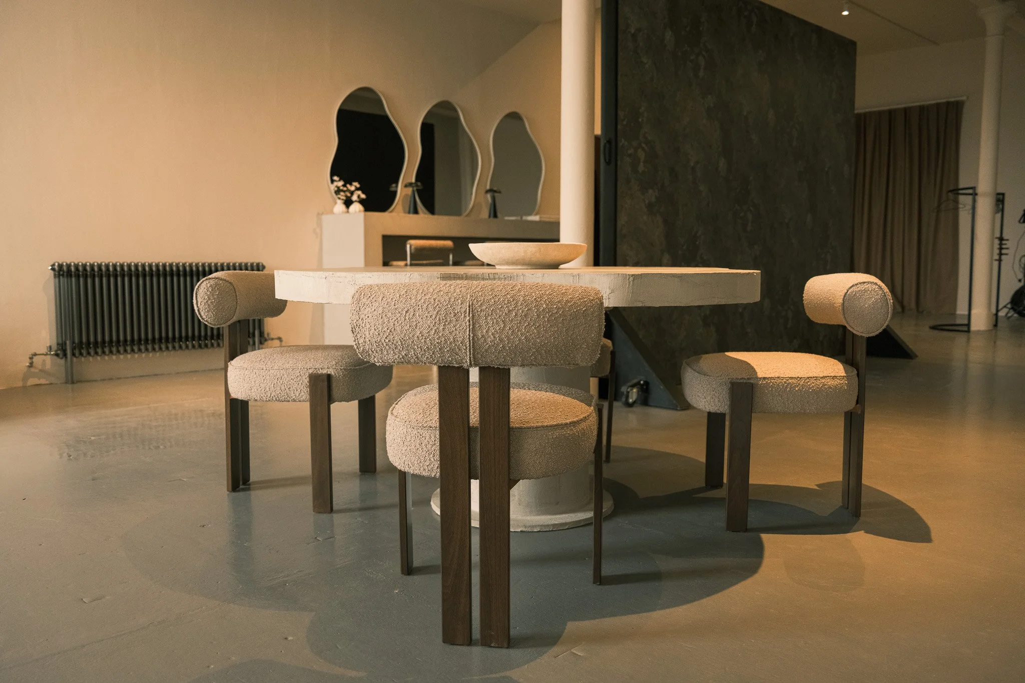 Interior of a modern room with a round marble table and four upholstered chairs with wooden legs and armrests, a radiator on the left, three mirrors on the wall, and a bowl on the table.