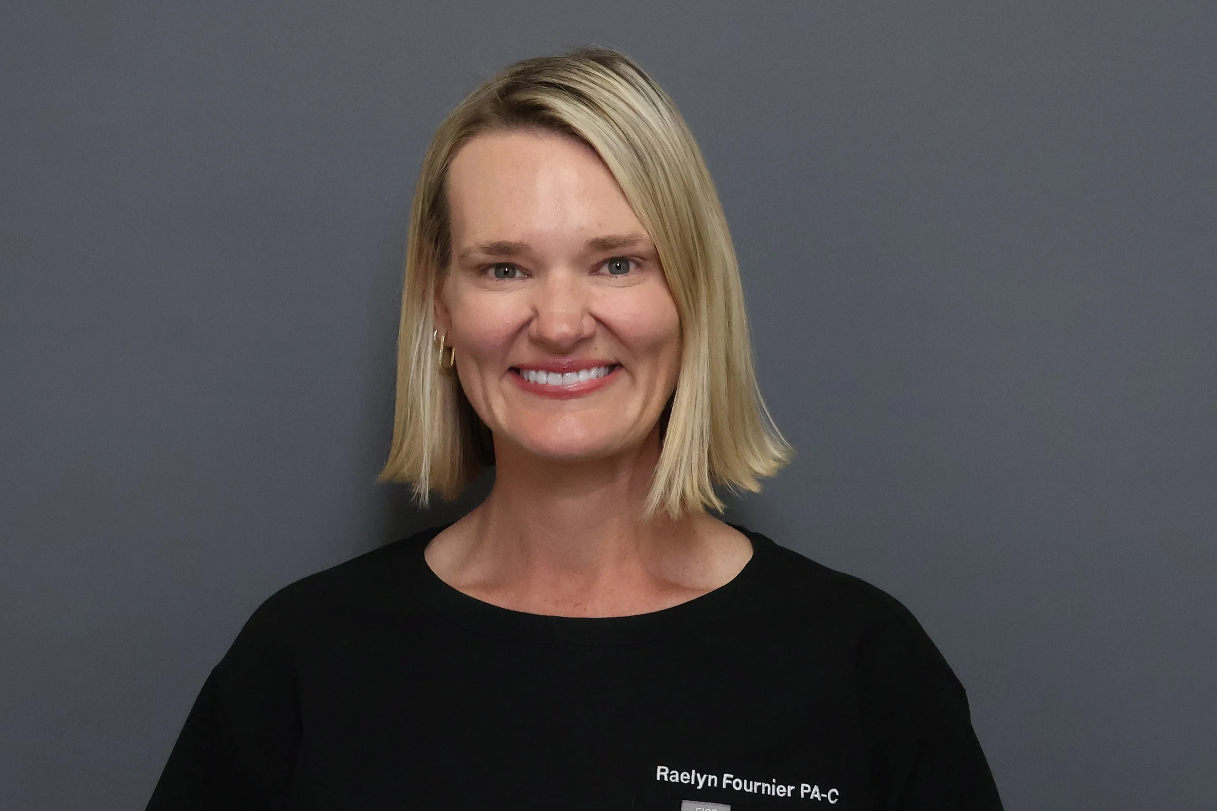 A smiling woman with shoulder-length blonde hair, wearing a black shirt with white text, standing against a gray background.