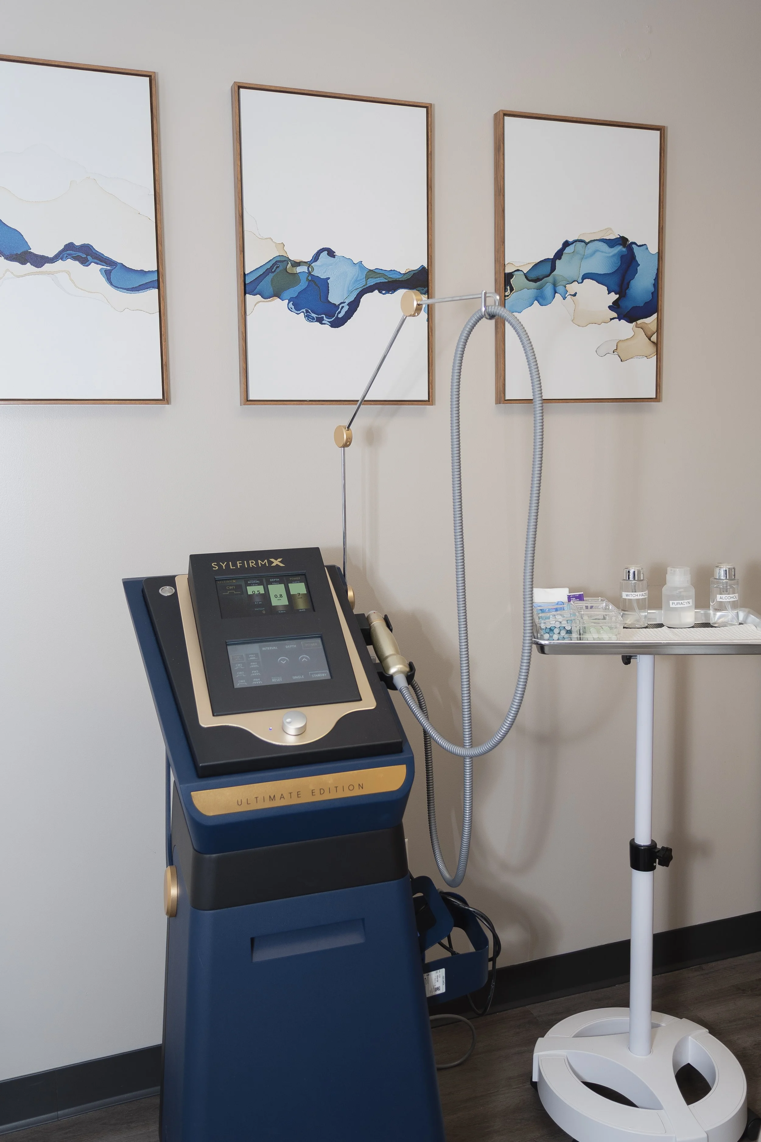 Medical laser or skincare device in a clinic room, with supplies on a nearby tray and modern abstract art on the wall.