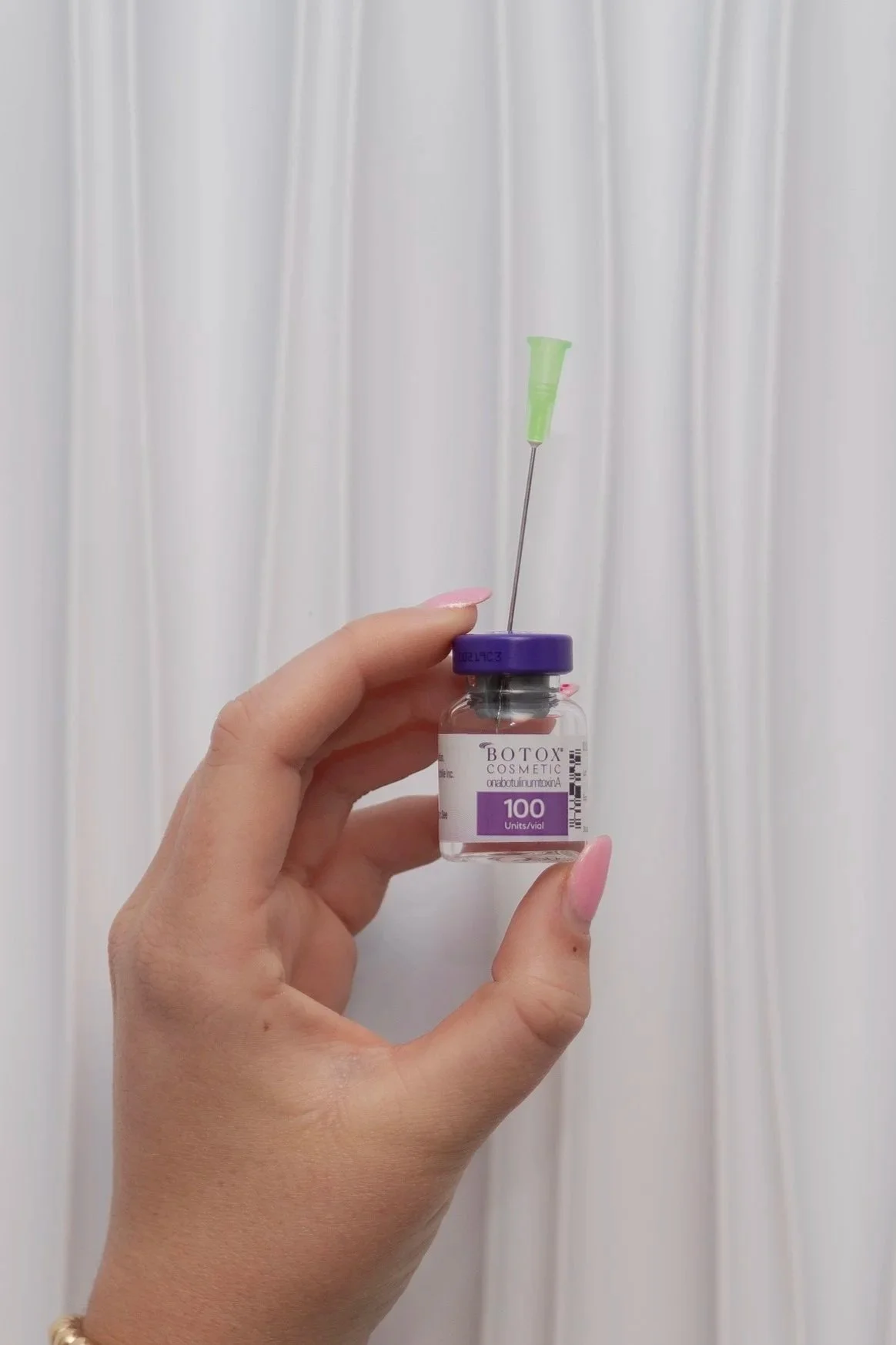 A hand holding a small vial labeled 'Botox Cosmetic,' with a syringe inserted into the vial against a white curtain background.