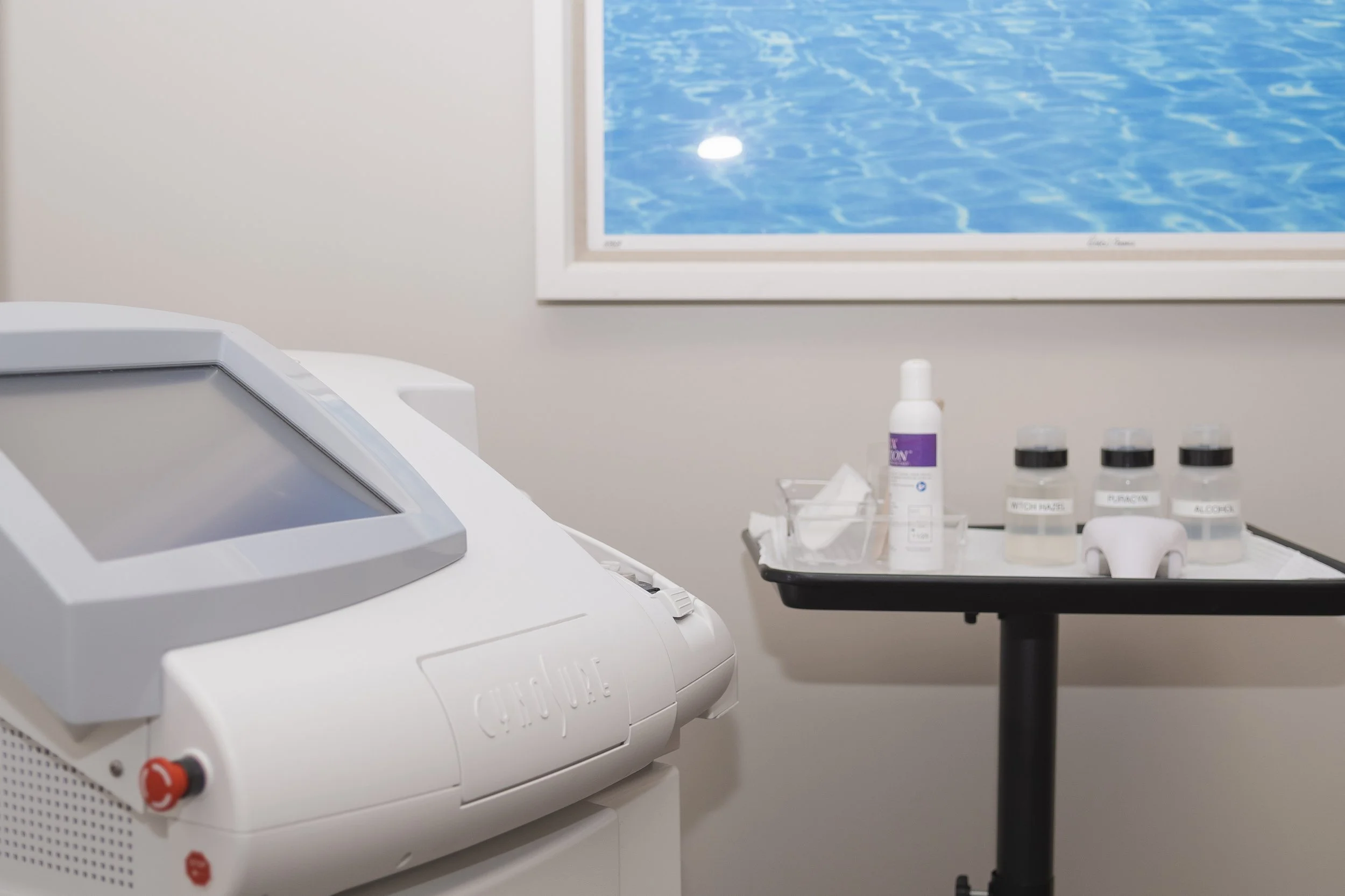 Medical equipment in a clinical setting including a white device with a touchscreen, a tray with bottles labeled 'Witch Hazel,' 'Platinum,' and 'Alcohol,' some tissues, and a framed picture of water on the wall.