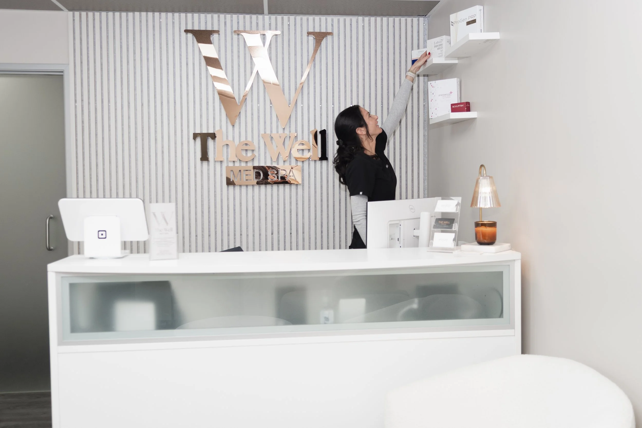 Reception area at The Well Medical Spa, with a woman reaching for products on a wall shelf behind the counter.