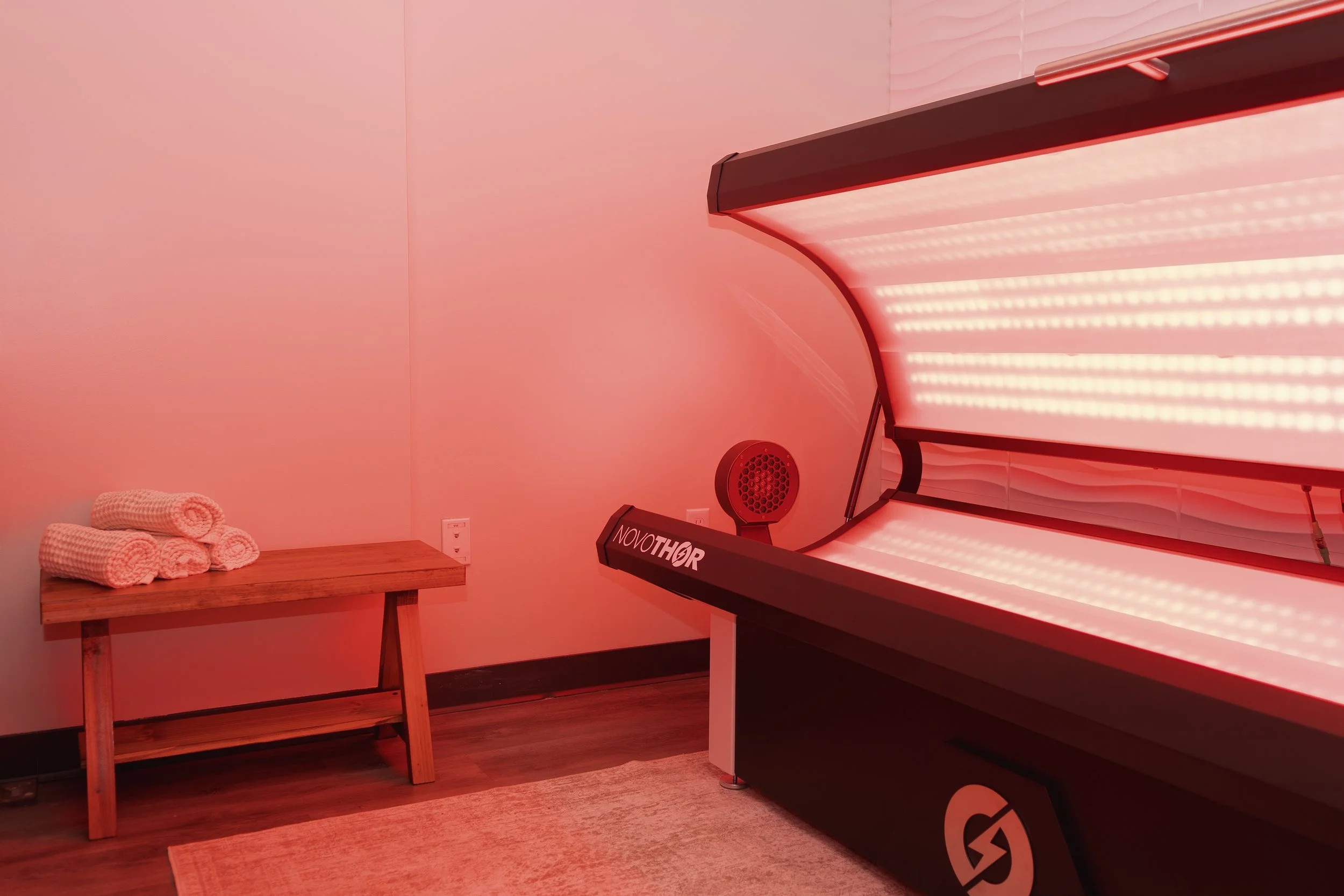 Indoor spa room with red light therapy bed, wooden bench with white rolled towels, and small speaker on the wall.