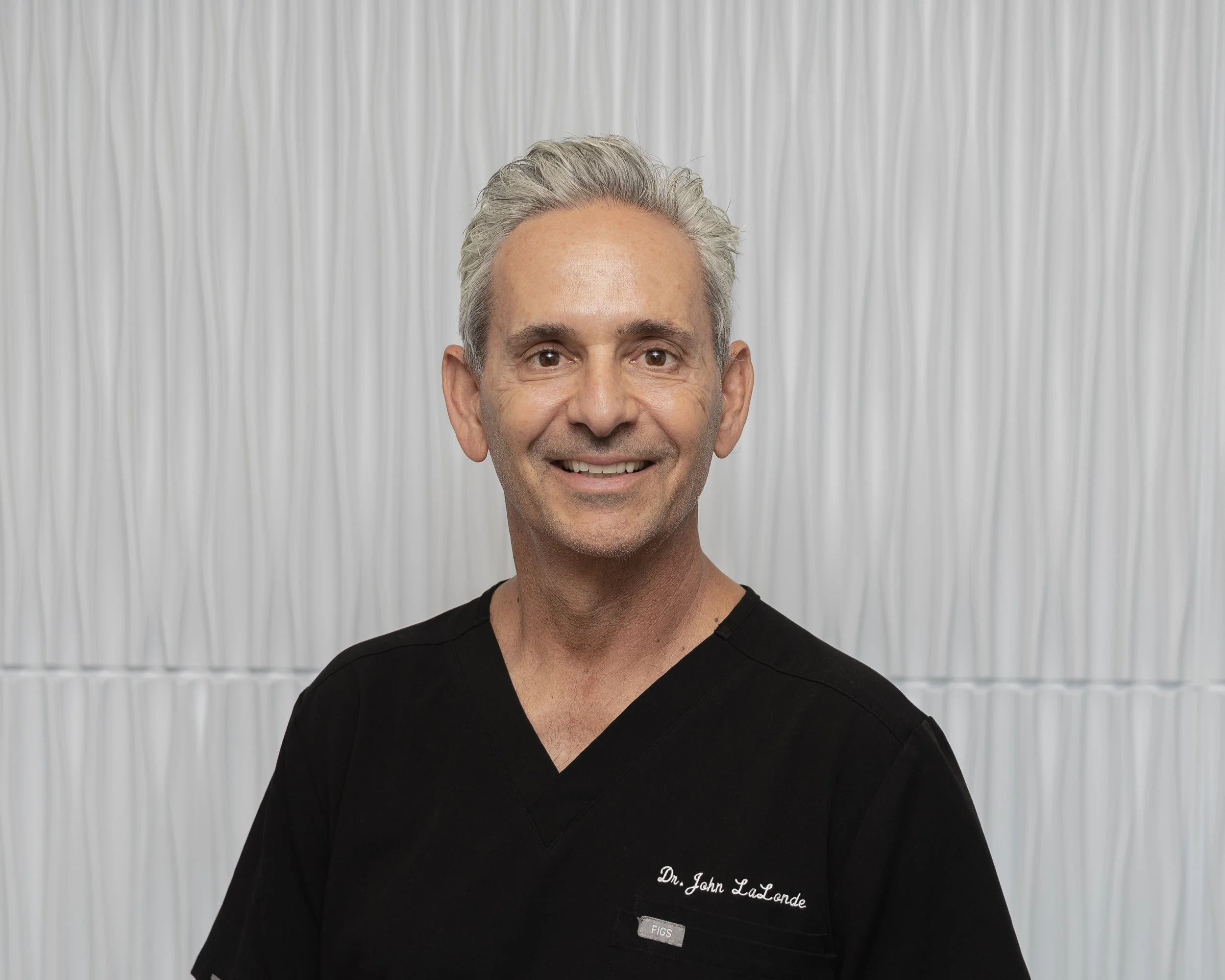 A middle-aged man with gray hair and a beard, wearing a white medical coat with the logo 'hoag,' smiling against a light gray background.