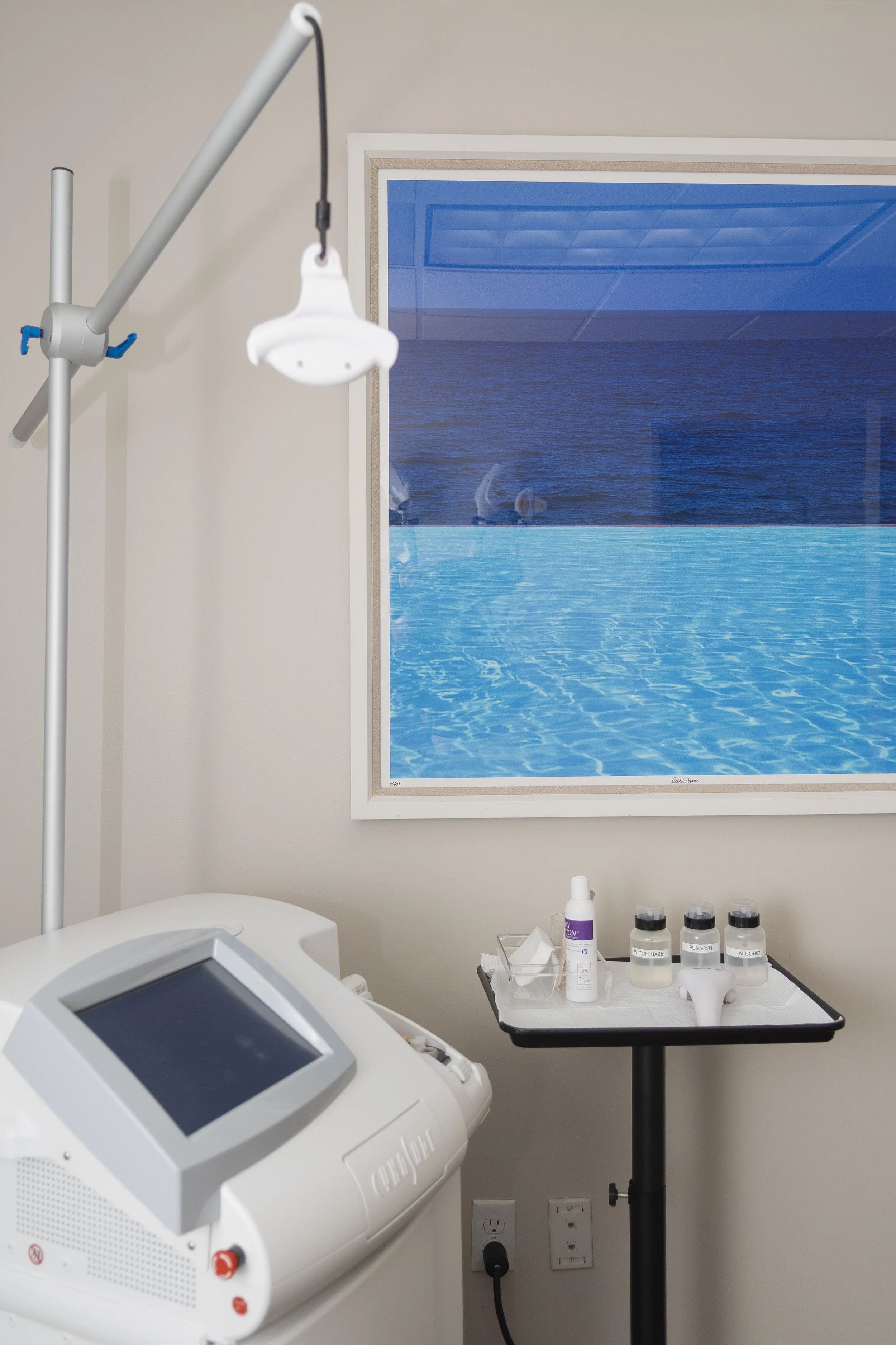Medical room with a machine, a small table with bottles and a spray, and a window showing a pool and ocean view.