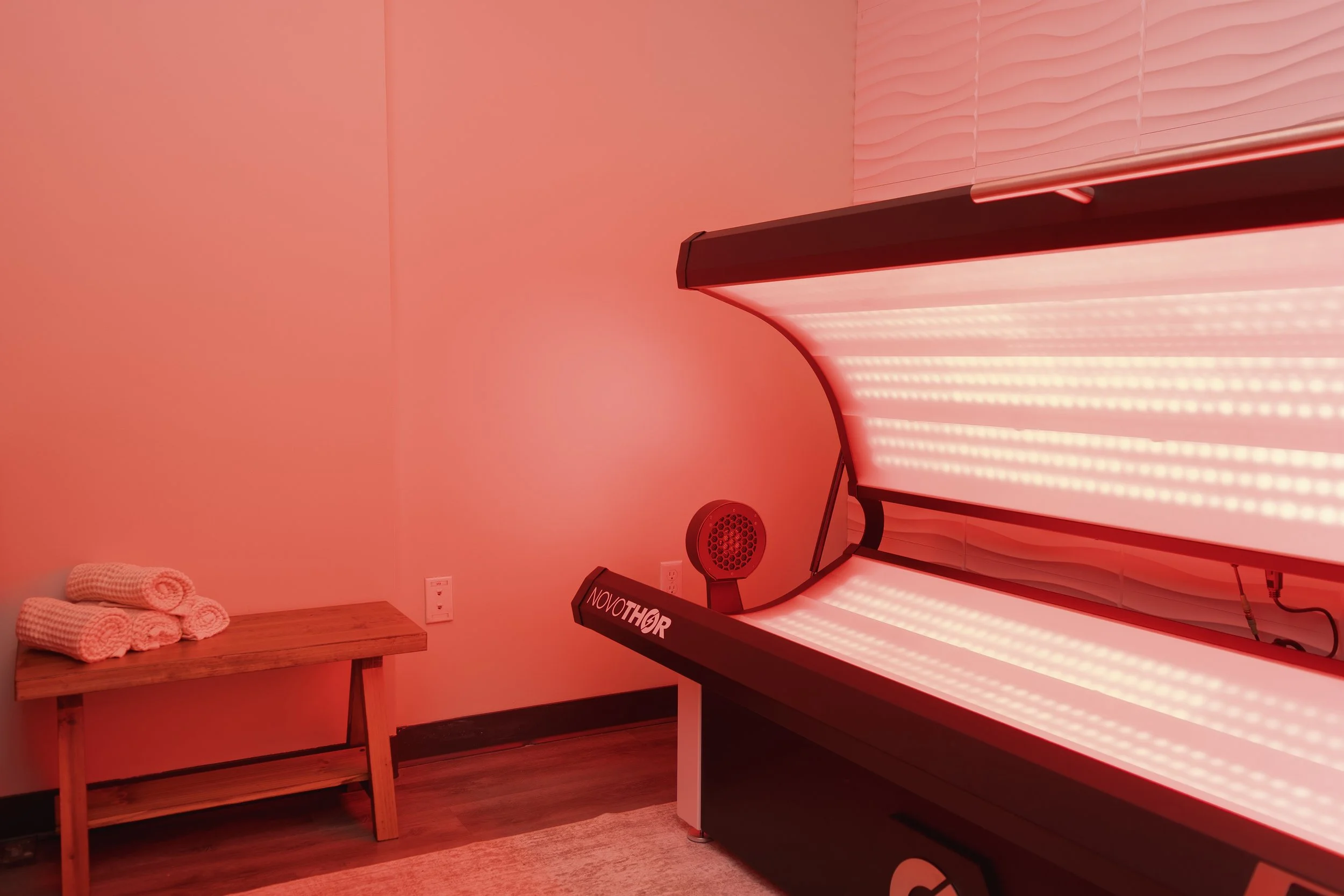 Indoor tanning bed illuminated in red light with a small wooden bench and three rolled towels on it, in a room with pinkish walls.