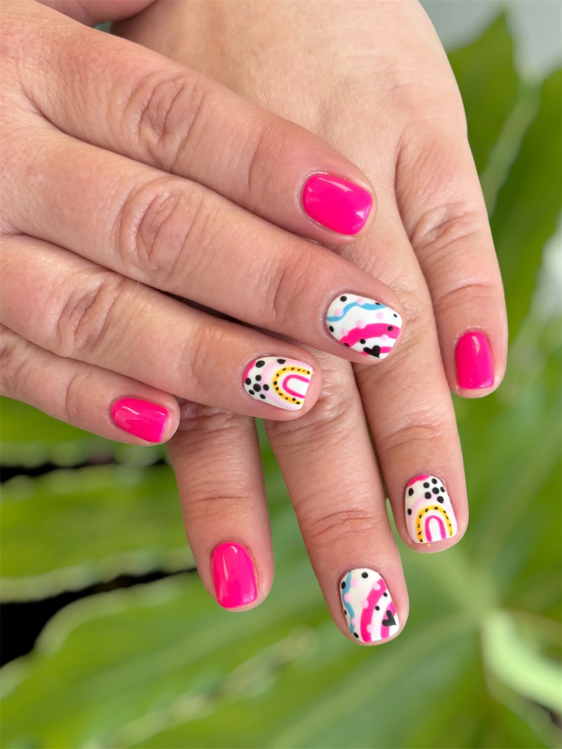 Close-up of a person's hand with pink painted fingernails and colorful, patterned nail art on some fingers, against a blurred green background.