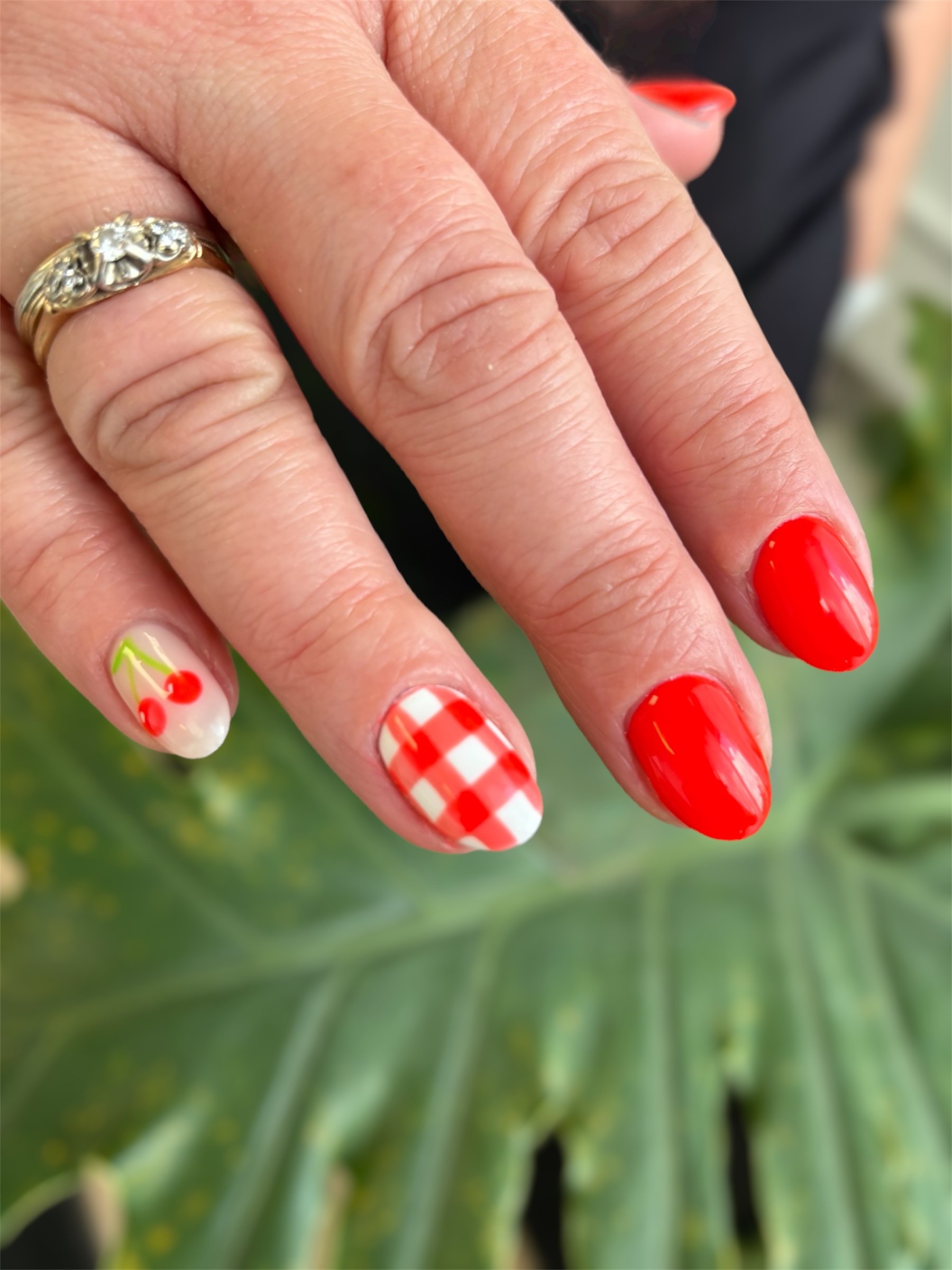 Close-up of a hand with colorful Christmas-themed nail polish and a gold ring with diamonds, in front of green foliage.
