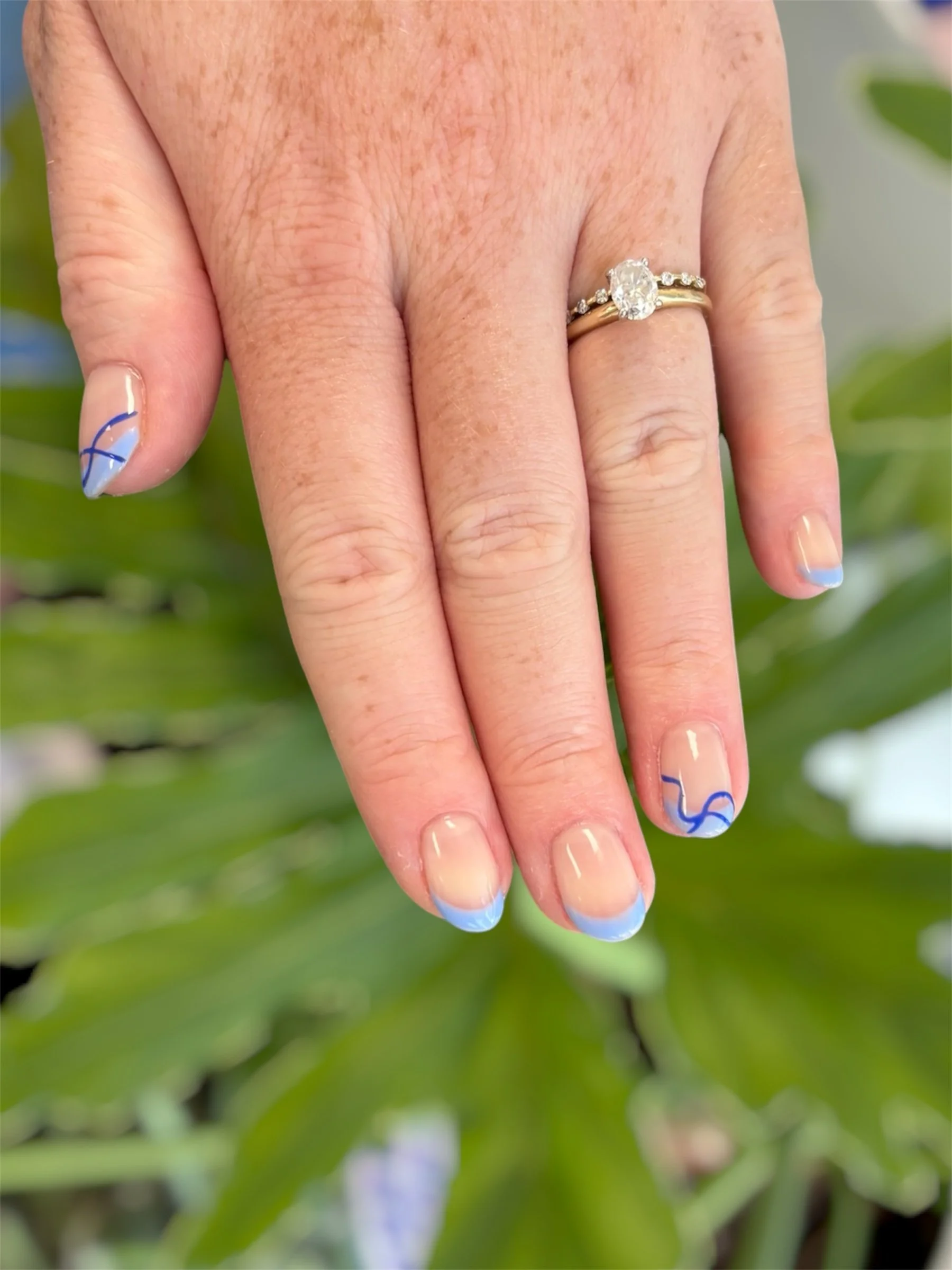 Close-up of a woman's hand displaying an engagement ring with a large central diamond and a wedding band with smaller diamonds. The nail polish is light blue with dark blue abstract design.