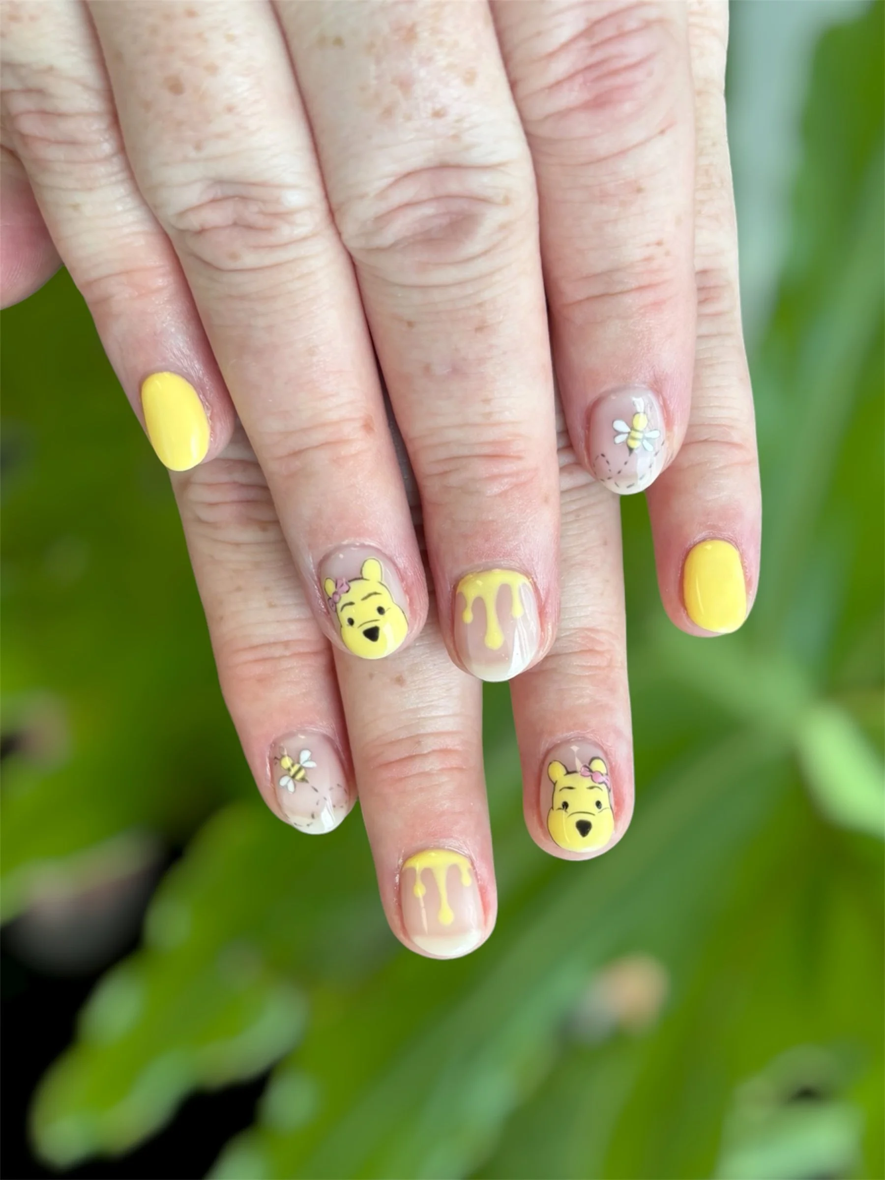 Close-up of a hand with yellow Winnie the Pooh themed nail art, including a character face, honey drips, bees, and flowers, with a green background.