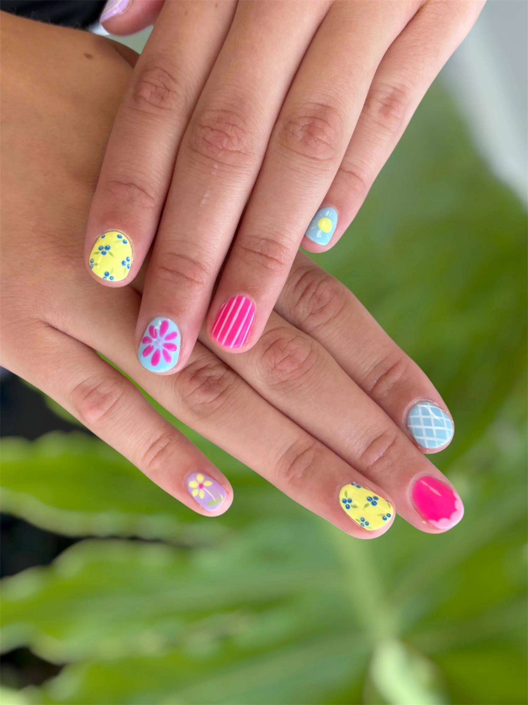 Close-up of fingers with colorful nail art featuring flowers, stripes, and patterns, against a blurred green background.