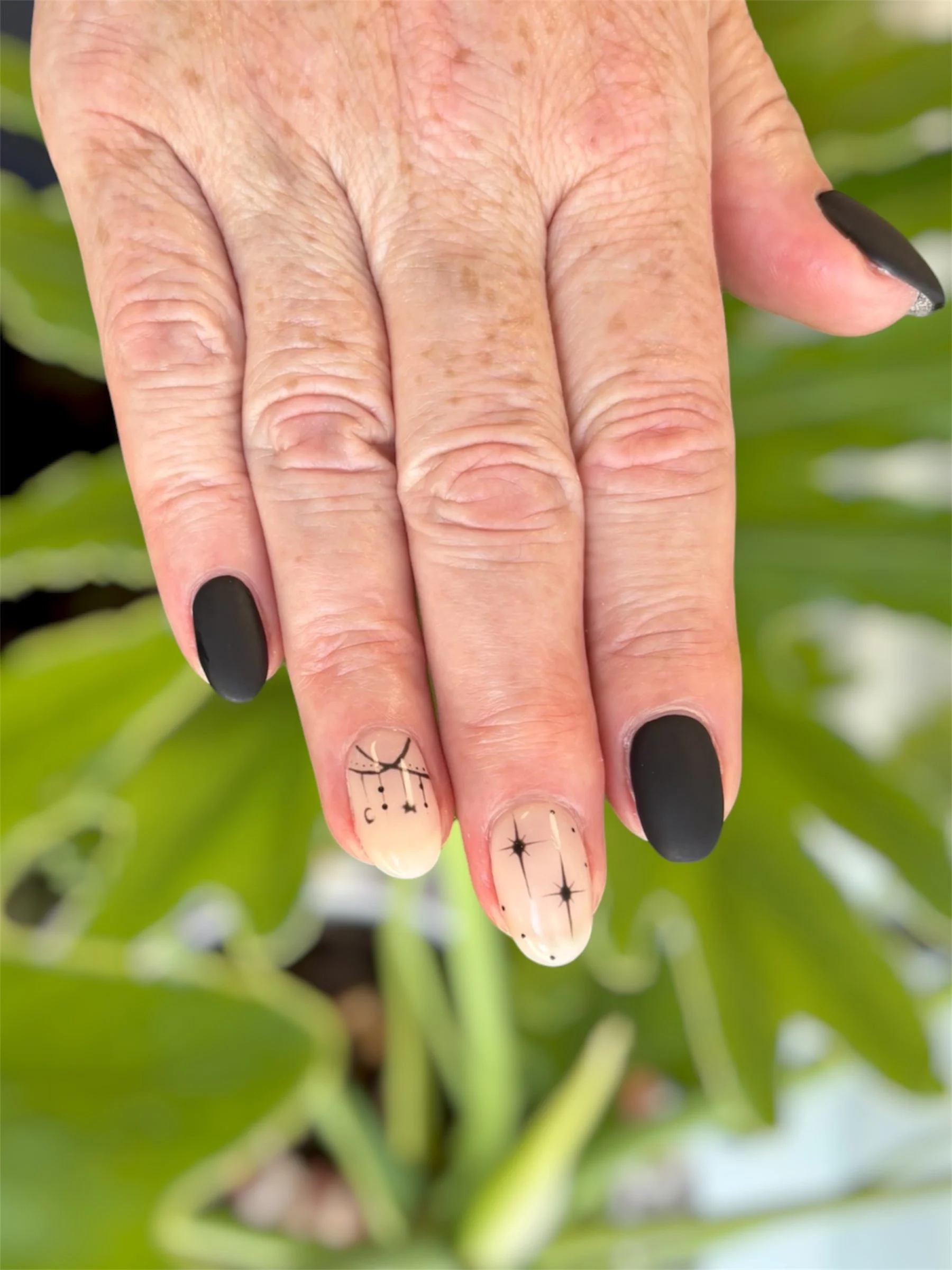 Close-up of a hand with black matte and decorated nails in front of a green leafy background.