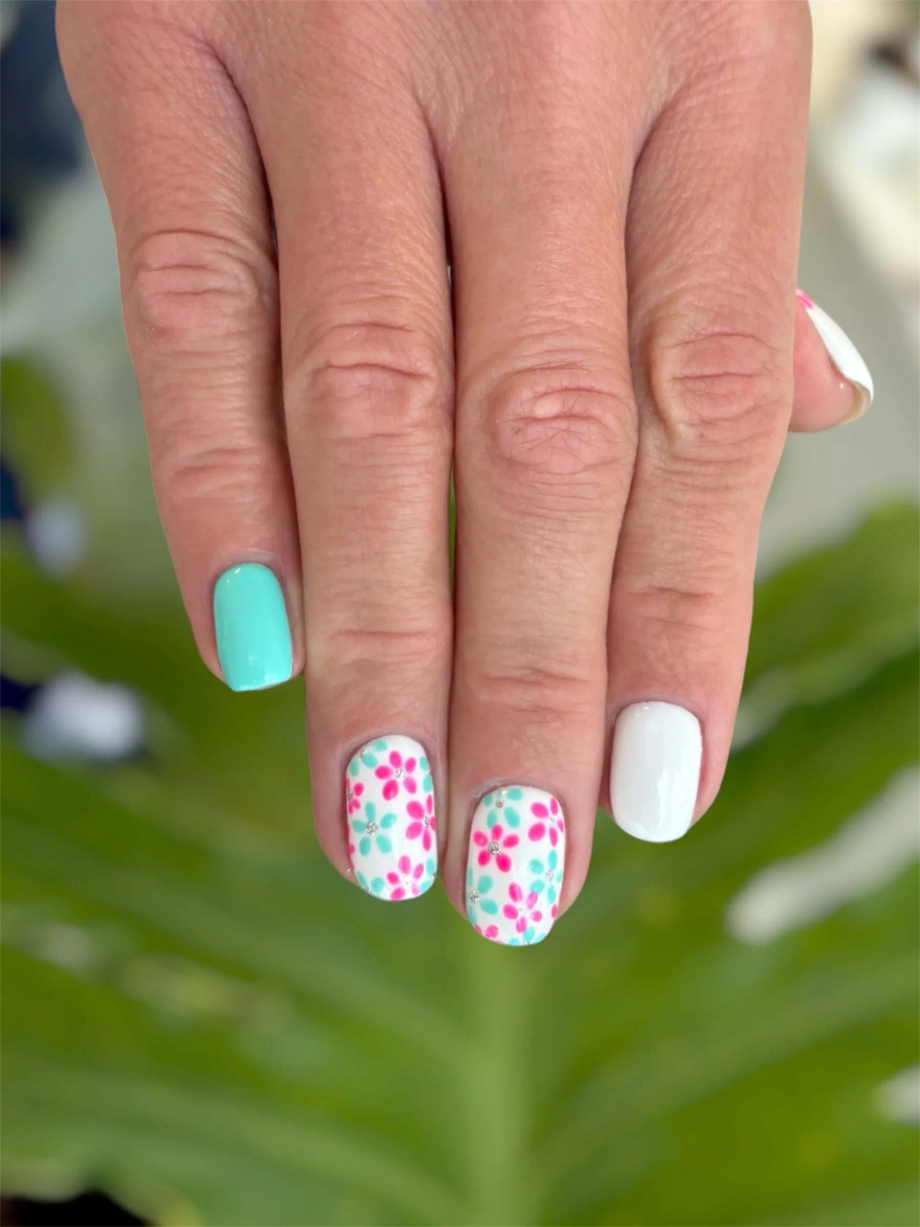 Close-up of a woman's hand with colorful nail art, including floral patterns and solid colors, set against a blurred green background.