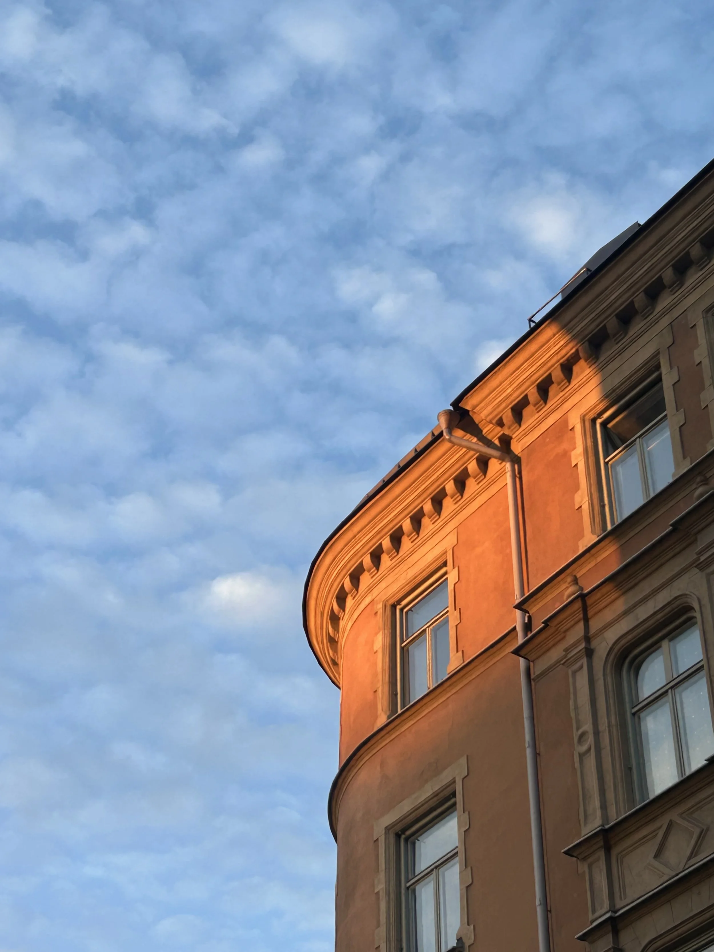 Edificio de ladrillo con ventanas y cornisa en la parte superior, iluminado por la luz del atardecer, con cielo azul con nubes alrededor.