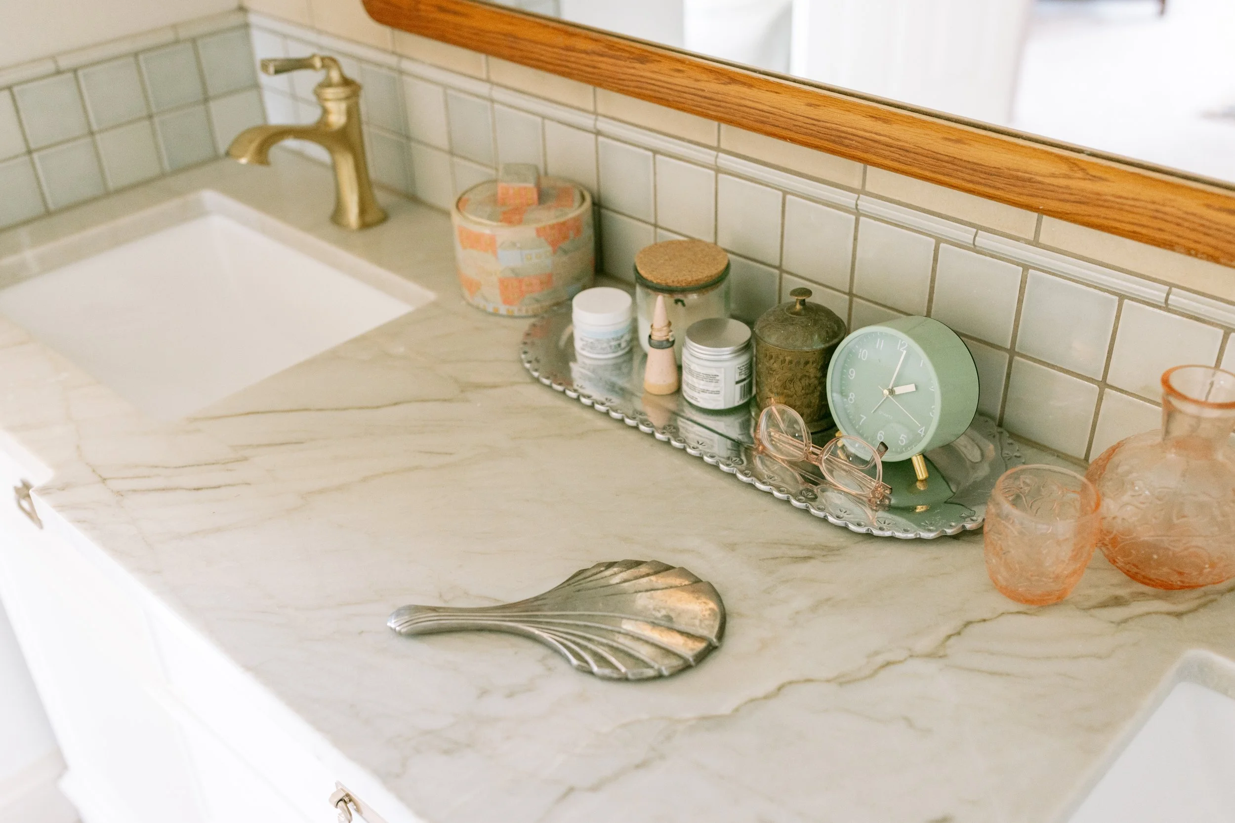 Bathroom countertop with various items including a silver tray, jars, a clock, and a pair of glasses in front of a tiled wall, with a sink and a faucet visible.