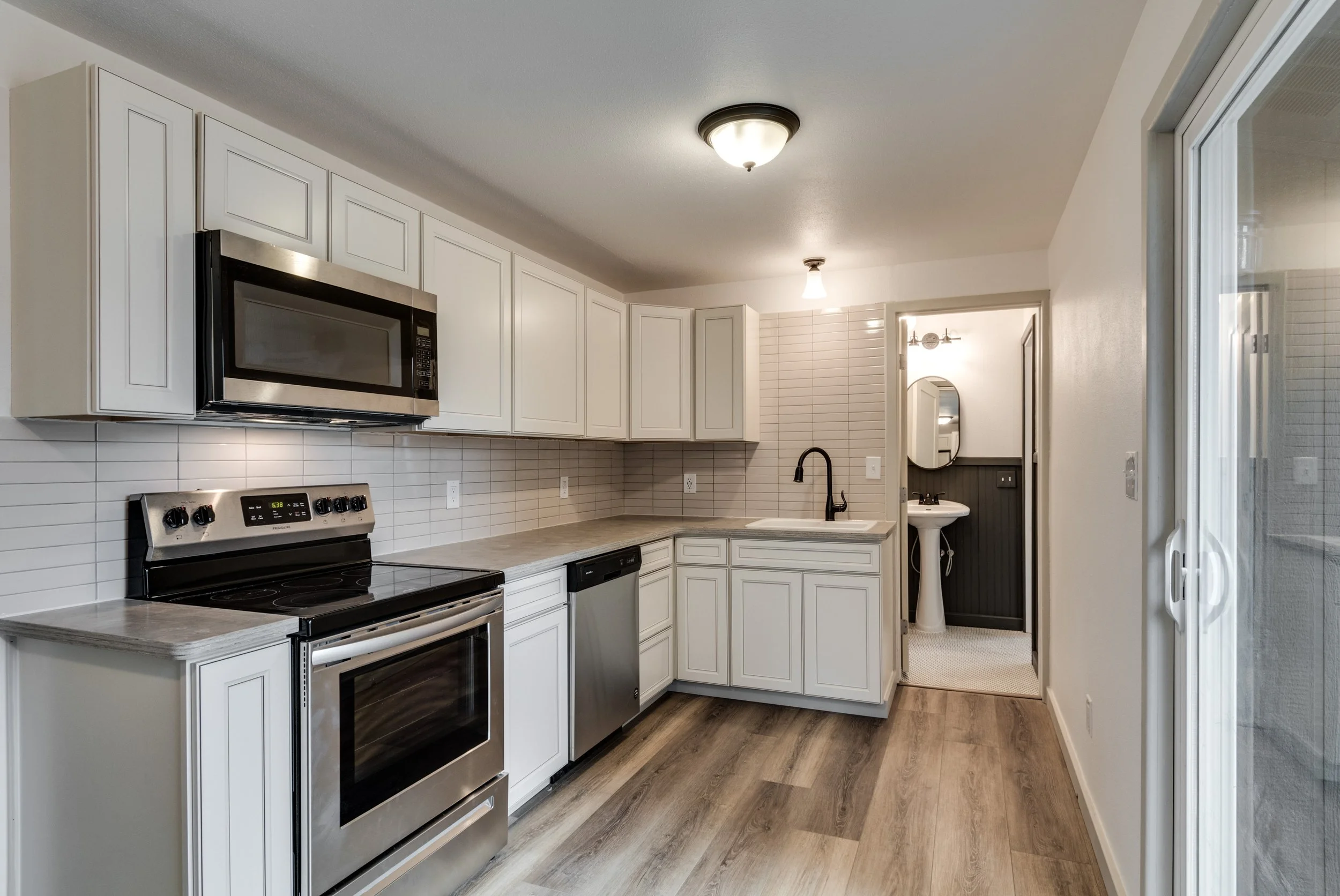 A kitchen with white cabinets, stainless steel appliances, a black faucet, and a small sink. There are hardwood floors and a doorway leading to a laundry area or bathroom.