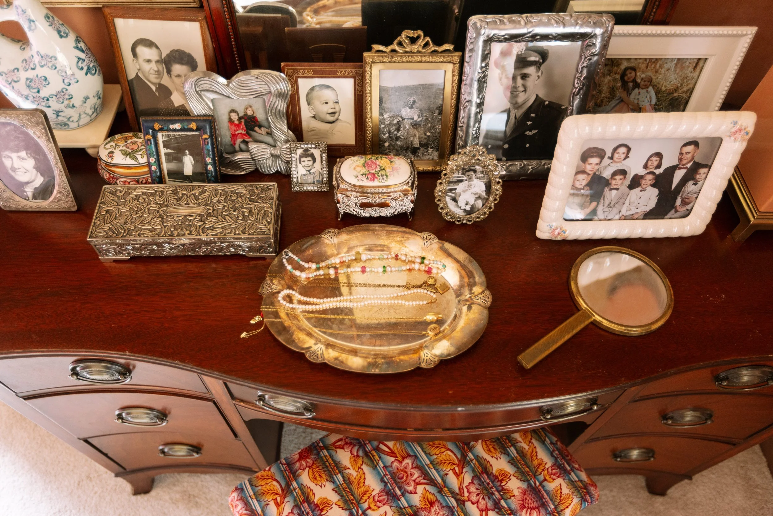 A wooden dresser with multiple vintage framed photographs, decorative boxes, jewelry, and a handheld mirror on top, along with a lampshade on the left and a patterned chair below.
