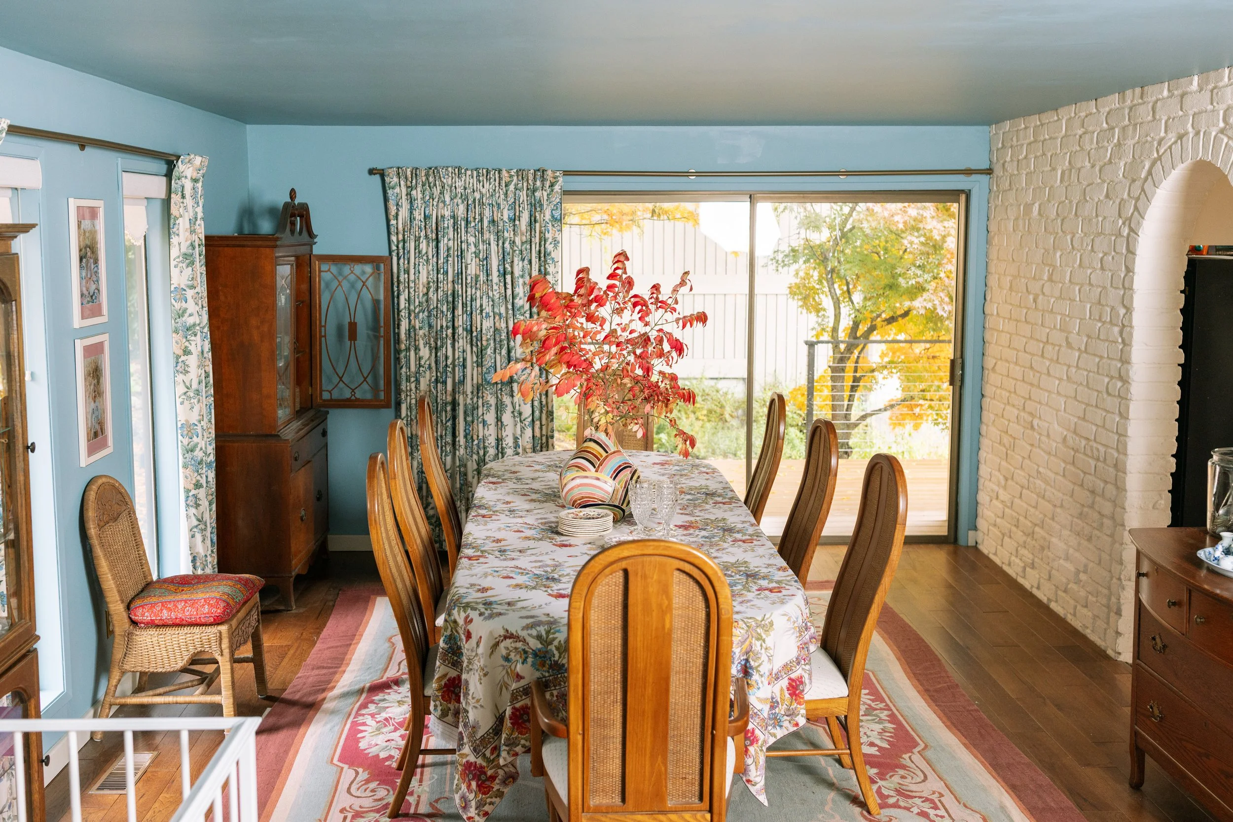A dining room with a large table covered with a floral tablecloth, surrounded by wooden chairs with woven backs. The table has glassware, plates, a striped pillow, and a floral centerpiece. The room has blue walls, floral curtains, a large window wit