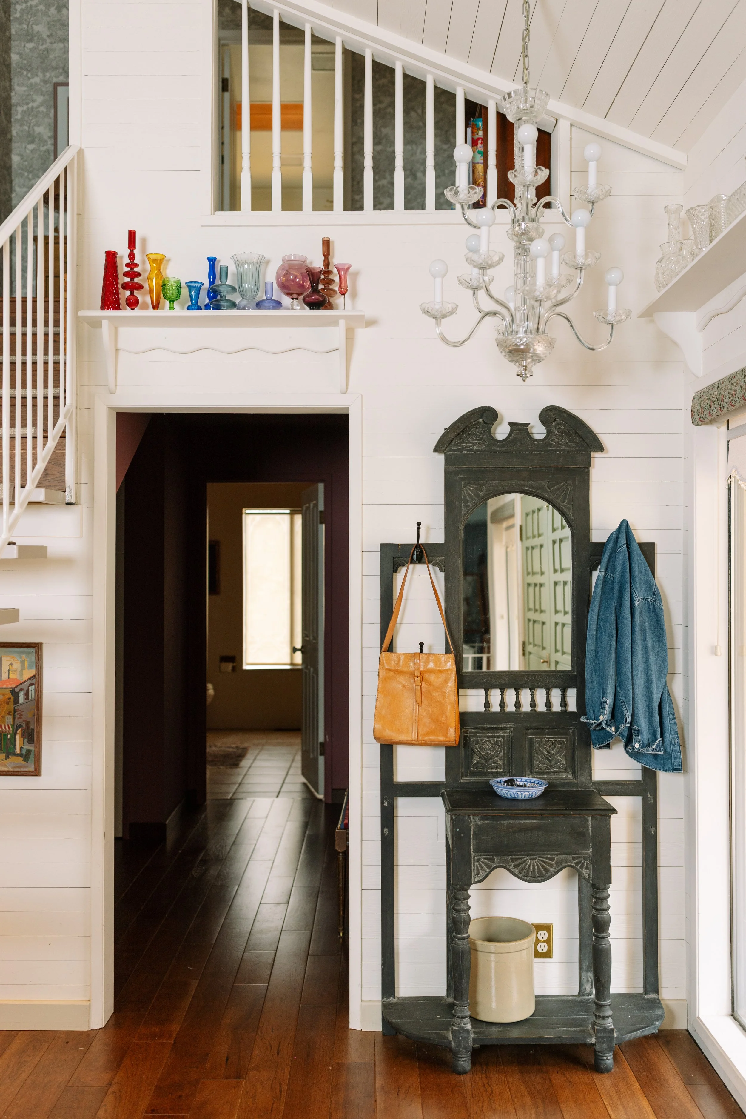 Entryway with antique black wooden hall tree holding a tan leather bag and a beige jar, a hanging coat, and a mirror on a white wall, chandelier, and colorful glass vases on a shelf.