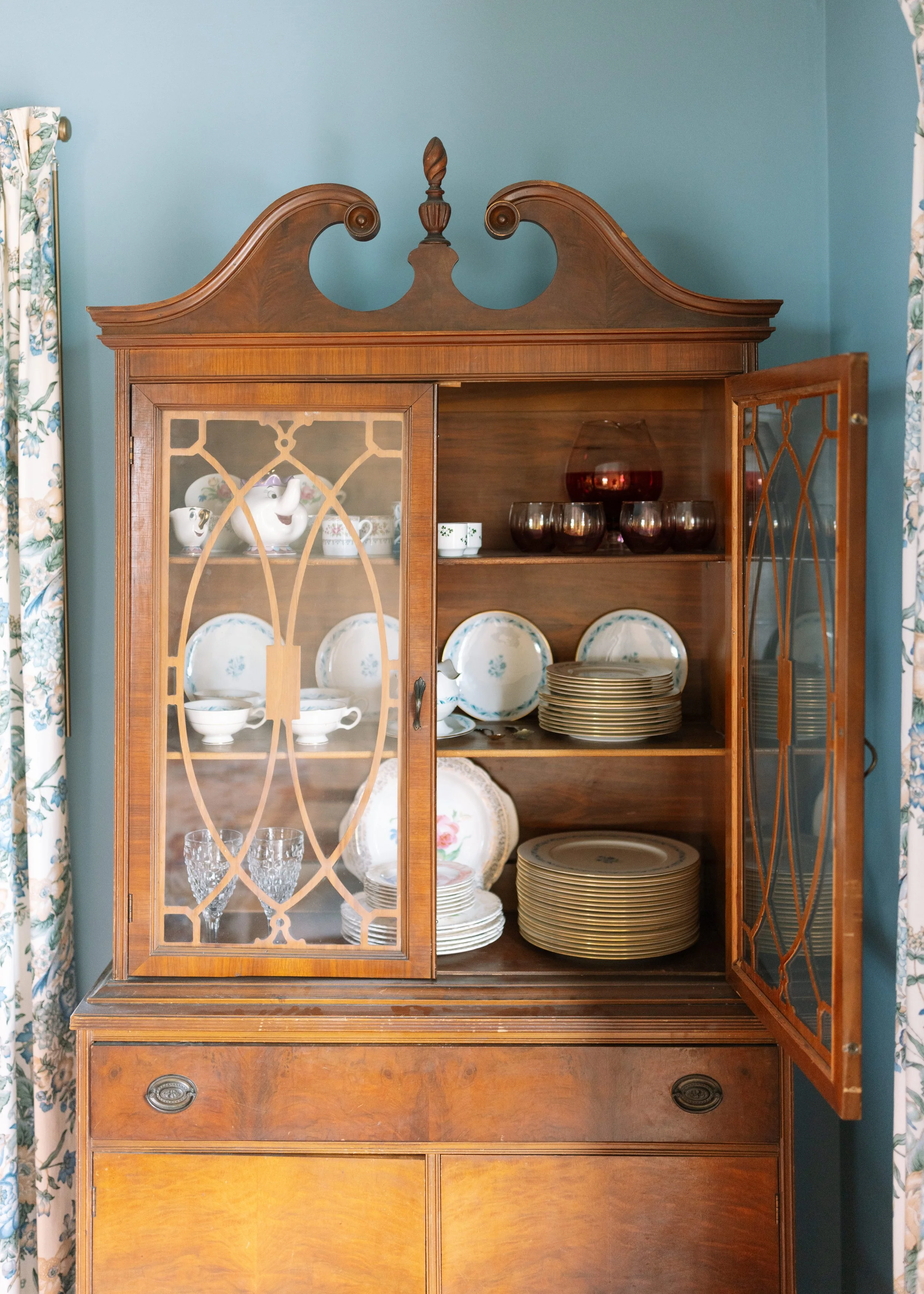 An antique wooden china cabinet with glass doors displaying teacups, bowls, and plates against a light blue wall with floral curtains.
