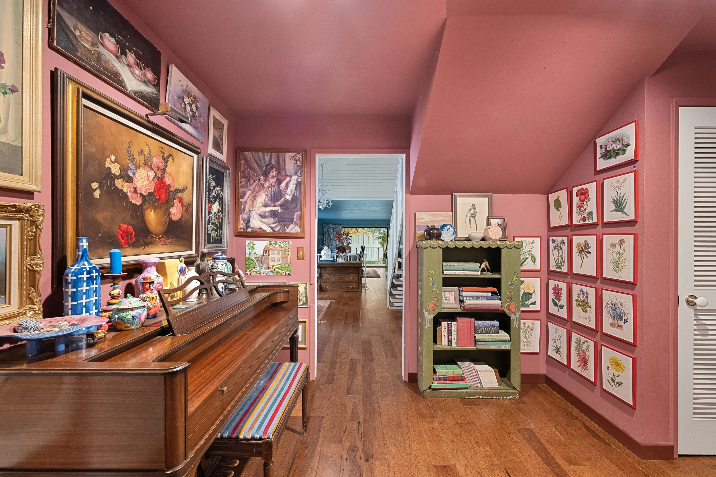 A cozy interior room decorated with paintings and framed botanical prints on pink walls. There is a wooden piano with decorative items on top, a colorful striped bench, and a green bookshelf filled with books and ornaments. An open doorway reveals a 