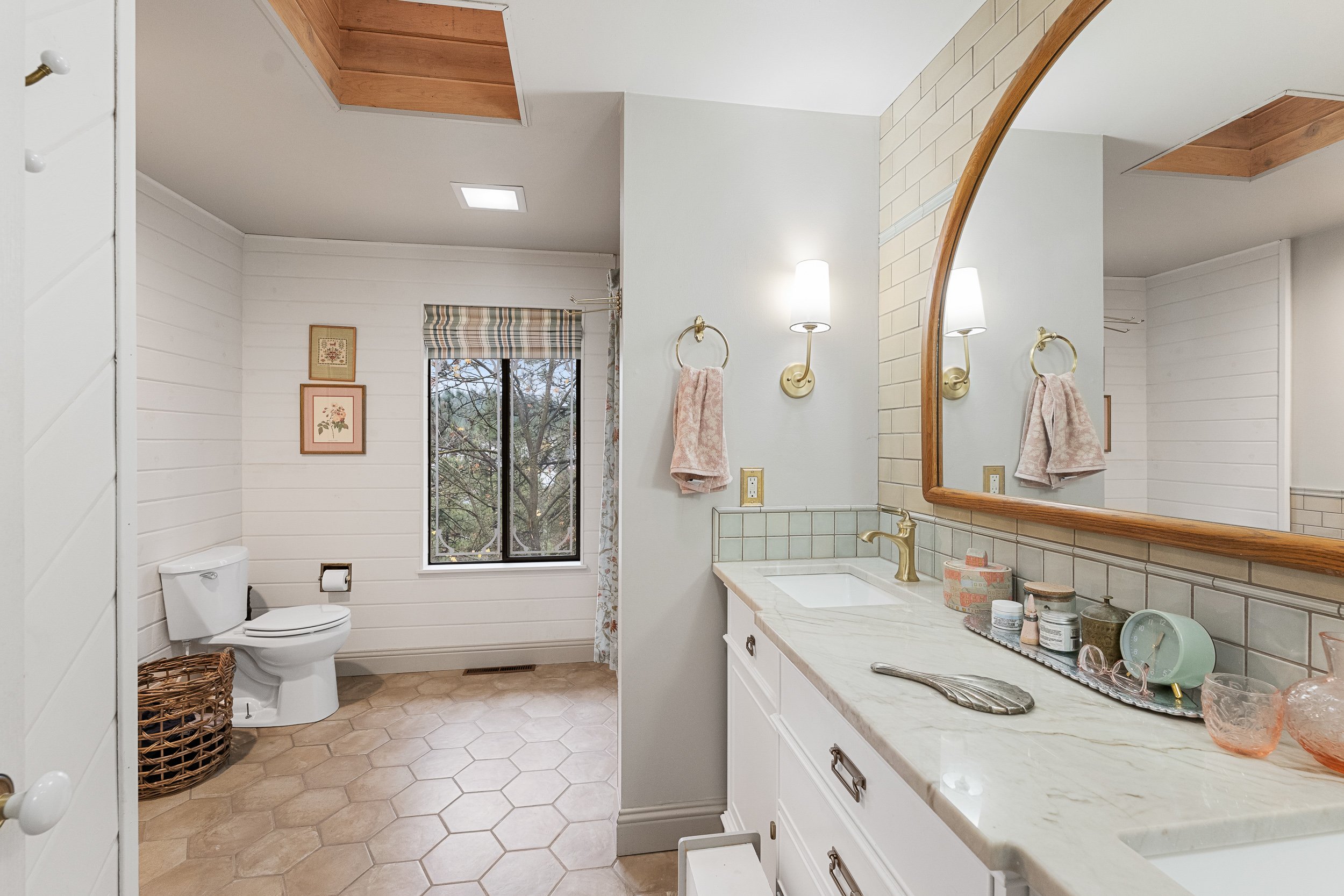 A bathroom with white shiplap walls, a window, and a white toilet. There is a wicker basket on the floor next to the toilet. The bathroom has a long white vanity with a marble countertop, a large wooden-framed mirror, and Brass wall-mounted lights. T