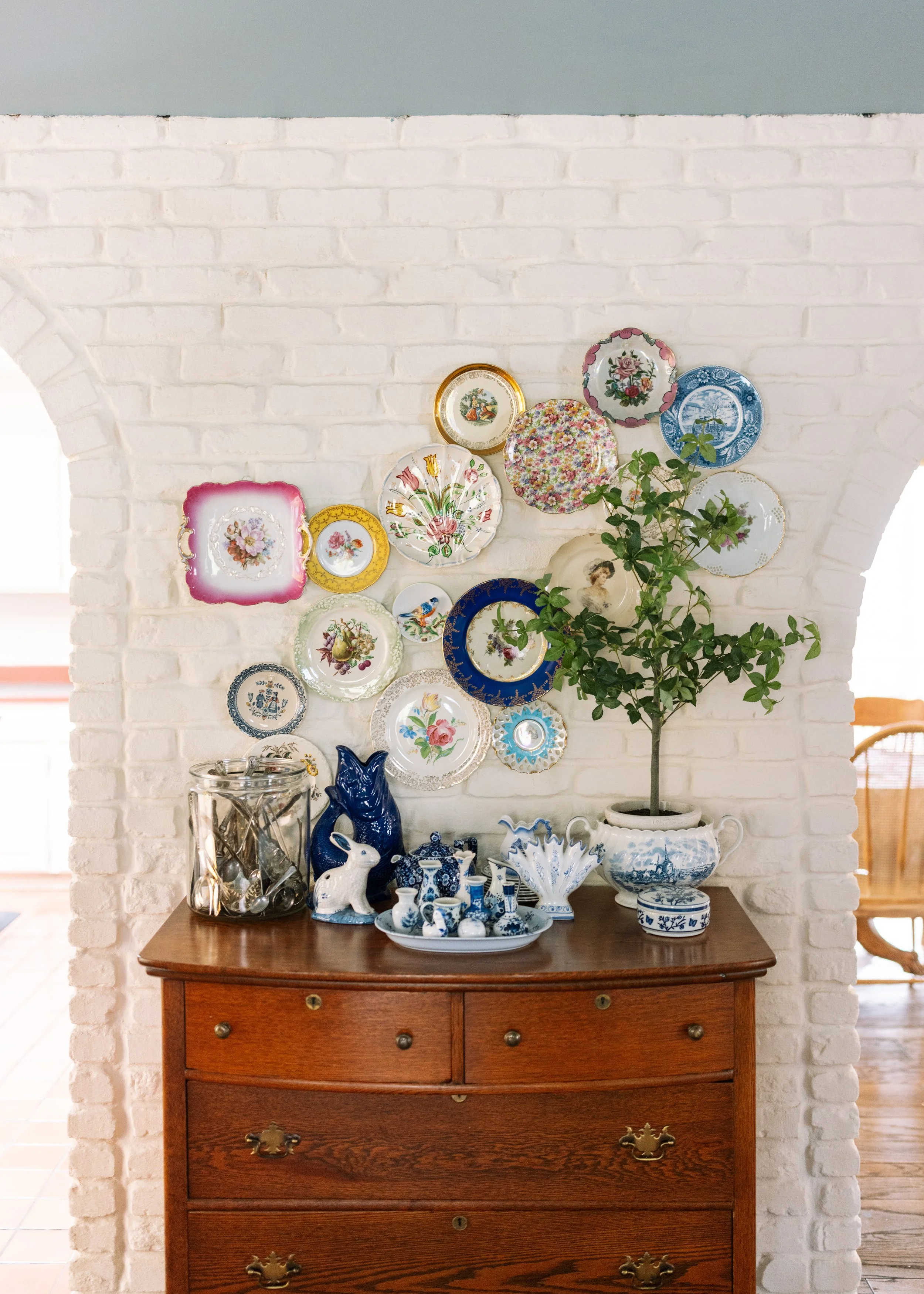 A wooden dresser with several drawers, topped with blue and white porcelain and ceramic figurines, including a rabbit and a dog, with a large ceramic bowl in the center. Above the dresser, a white brick wall displays a collection of decorative plates