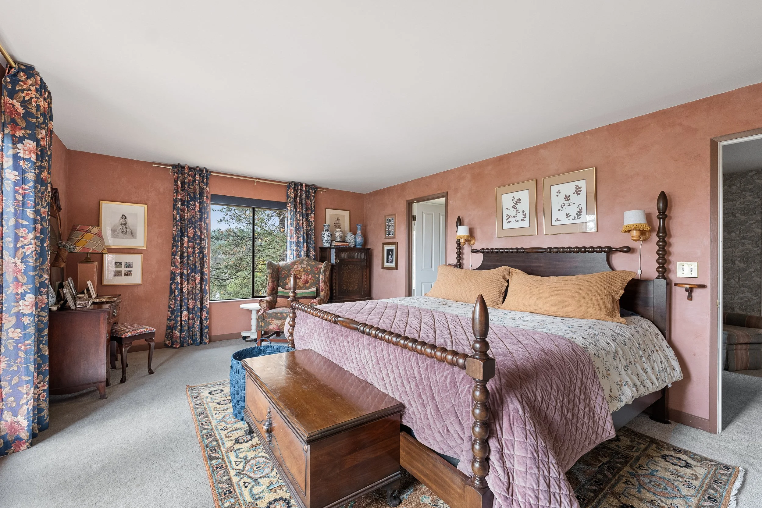 A bedroom with a large wooden bed, pink quilt, a wooden chest at the foot, patterned curtains, a floral armchair, multiple framed pictures on the wall, and a window showing trees outside.