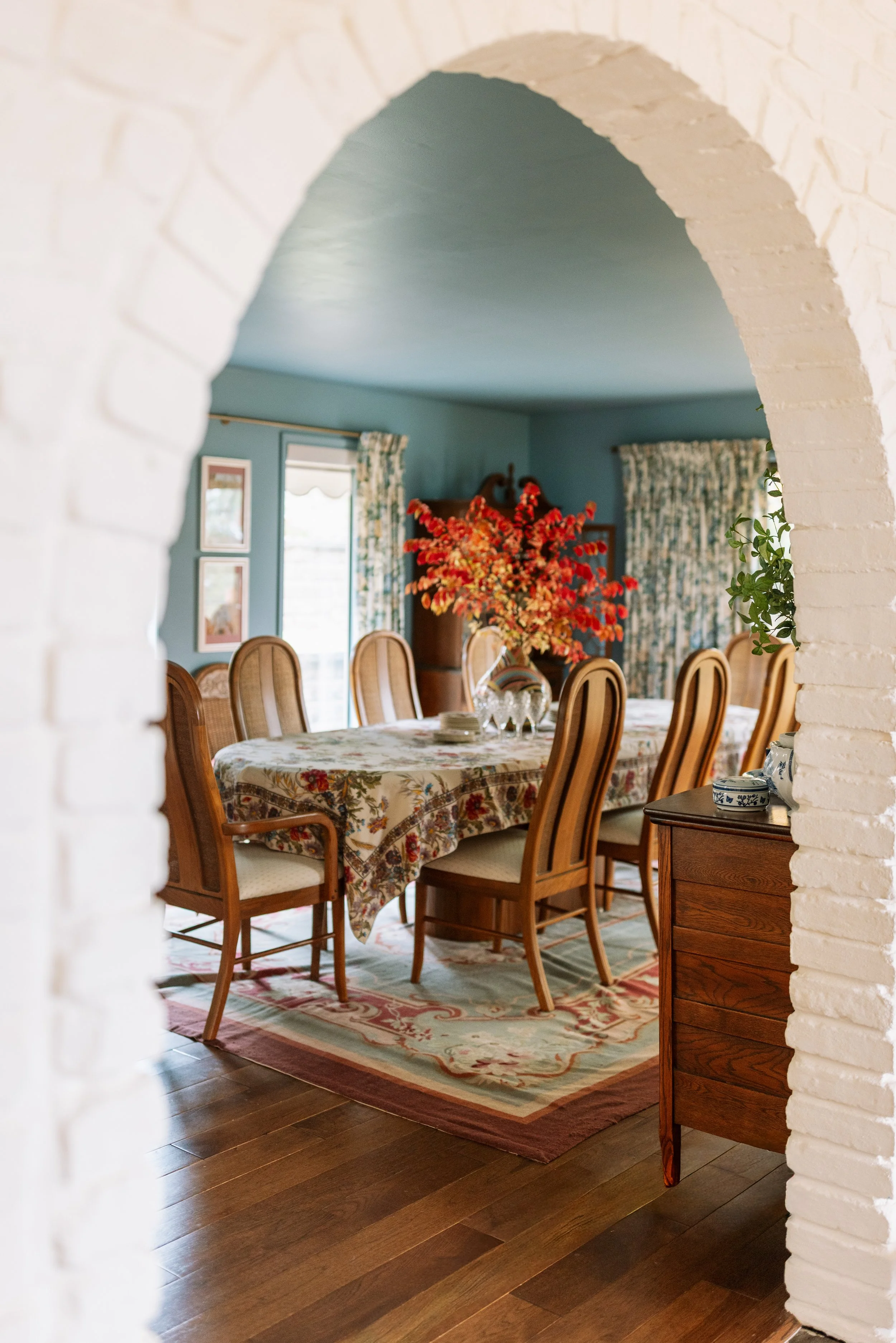 A dining room with a long wooden table covered by a floral tablecloth, surrounded by wooden chairs with woven rattan backs. There is a large floral arrangement in a vase on the table, and a sideboard with a small decorative dish. The room has blue wa