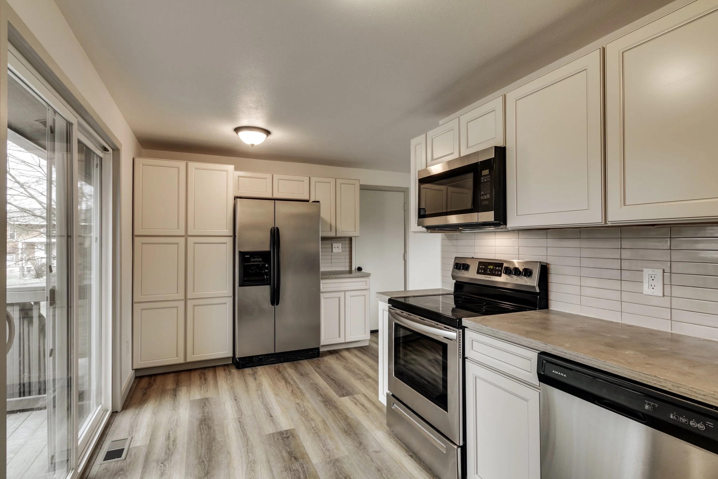 A bright modern kitchen with white cabinets, stainless steel appliances including a refrigerator, microwave, and stove, light wood flooring, and a sliding glass door leading outside.