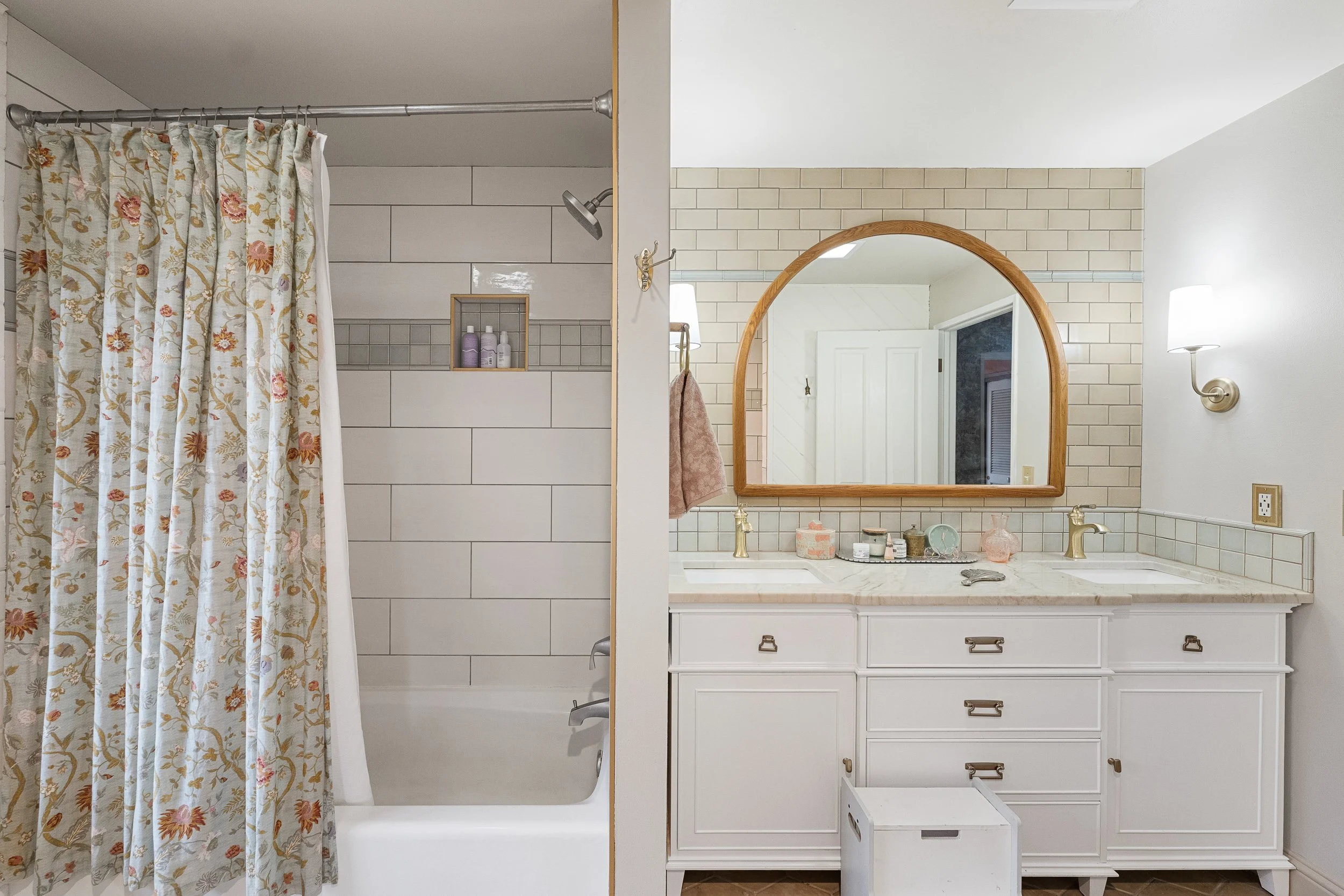 A bathroom with a shower area on the left with floral patterned curtain and a vanity with a large mirror on the right, decorated with a soap dispenser, jars, and a small tray. The bathroom has white furniture, beige tiles, and wall-mounted lamps.