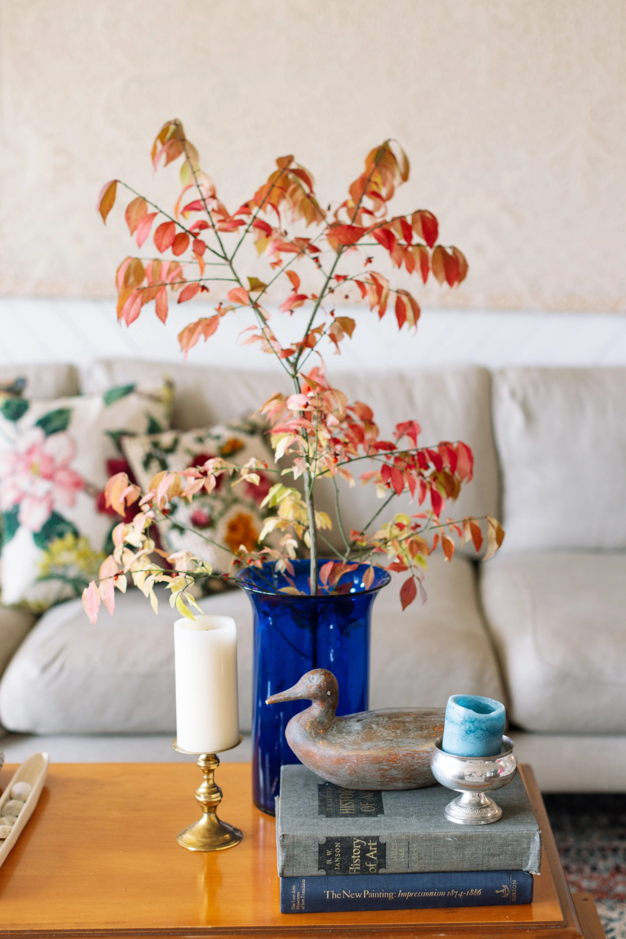 Decorative table display with a blue vase holding a branch with red and orange leaves, surrounded by a cream-colored candle on a gold holder, a ceramic duck sculpture, and three art history books on a wooden table.