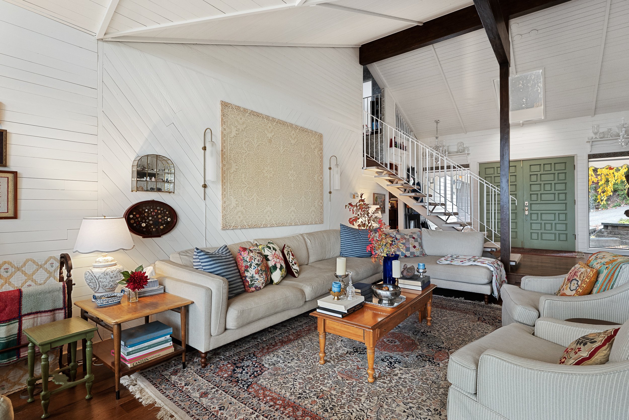 Living room with white paneled walls, a beige sectional sofa with colorful pillows, a wooden coffee table with books and candles, a patterned area rug, and a staircase leading to an upper level. Decor items include a wall mirror, lamps, and framed pi