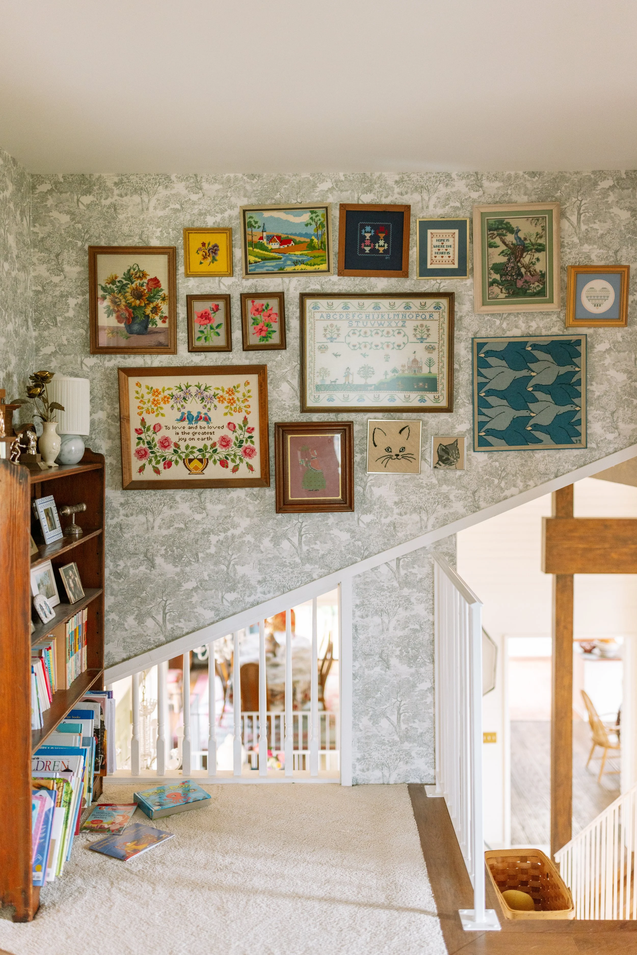 View of a staircase landing with wall decorations, including framed cross-stitch art, paintings of flowers and landscapes, and small prints, with a bookshelf filled with books on the left and a view into a living or dining area below.