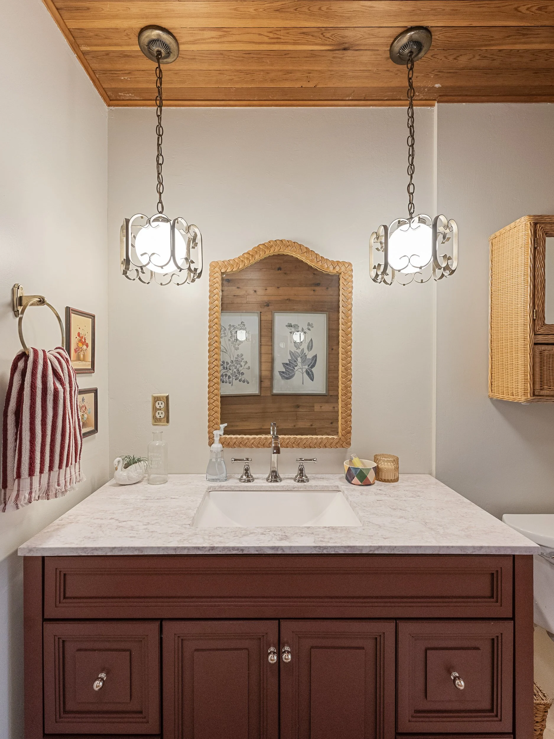 Bathroom sink with a brown cabinet, marble countertop, decorative mirror with woven frame, and two modern pendant lights with chain links hanging from a wooden ceiling.
