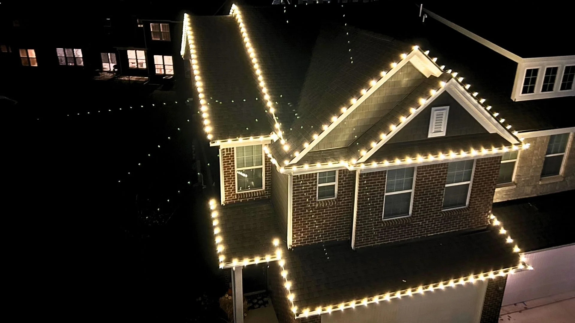 Nighttime aerial view of a brick house decorated with string lights outlining the roof and roof peak.