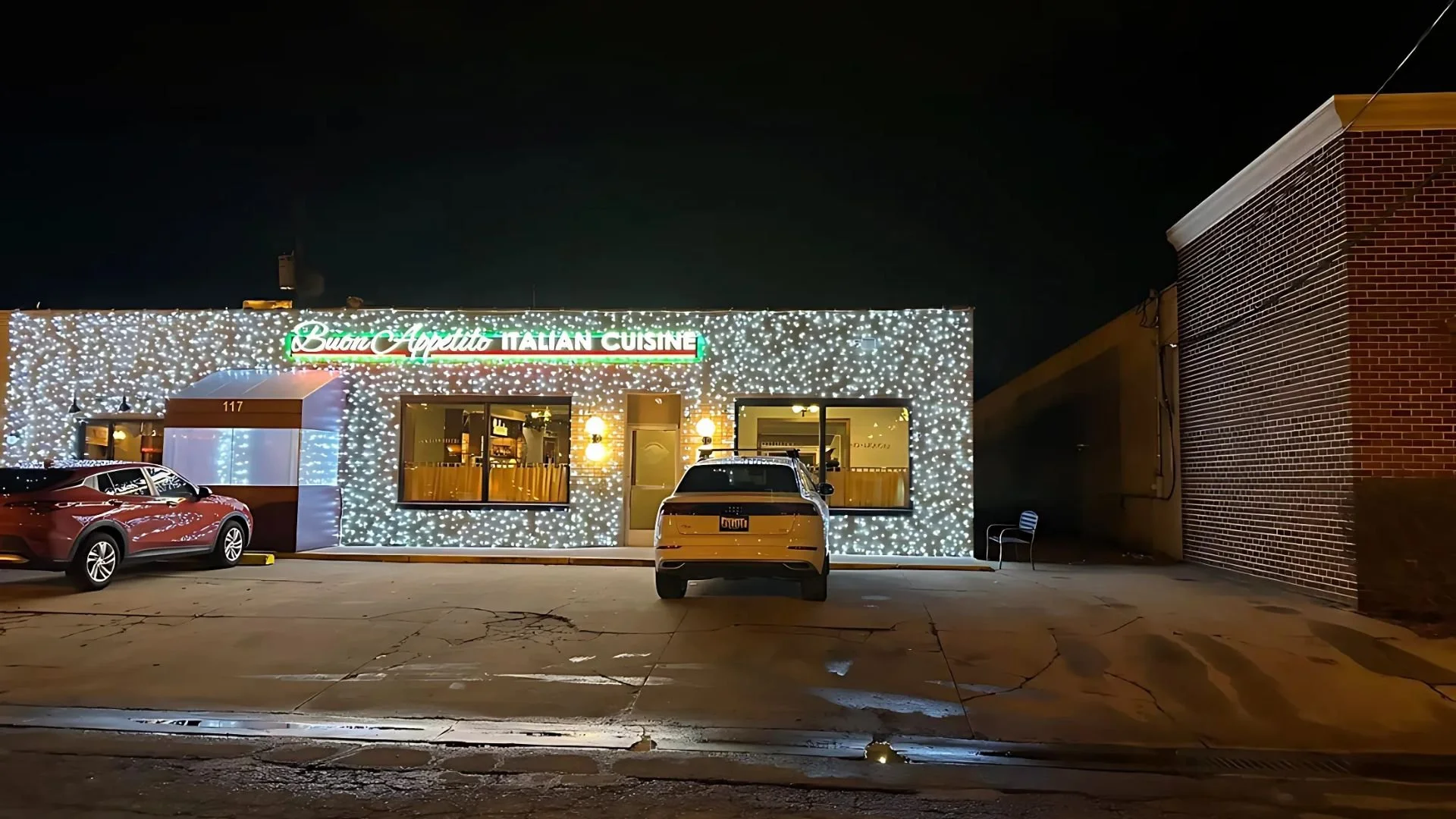 A restaurant with a sign that reads 'Buona Vità Italian Cuisine' decorated with white string lights outside at night. There are three cars parked in front, with the middle one facing directly towards the camera, and a brick wall on the right side.