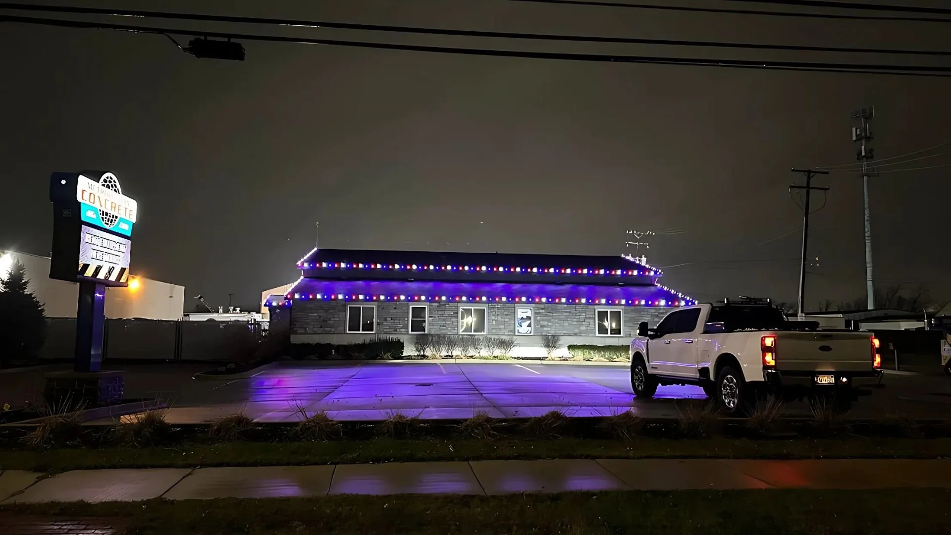 Nighttime street scene with a building decorated with purple and white string lights, a white pickup truck parked in front, and a sign on the left side reading 'Construct'.